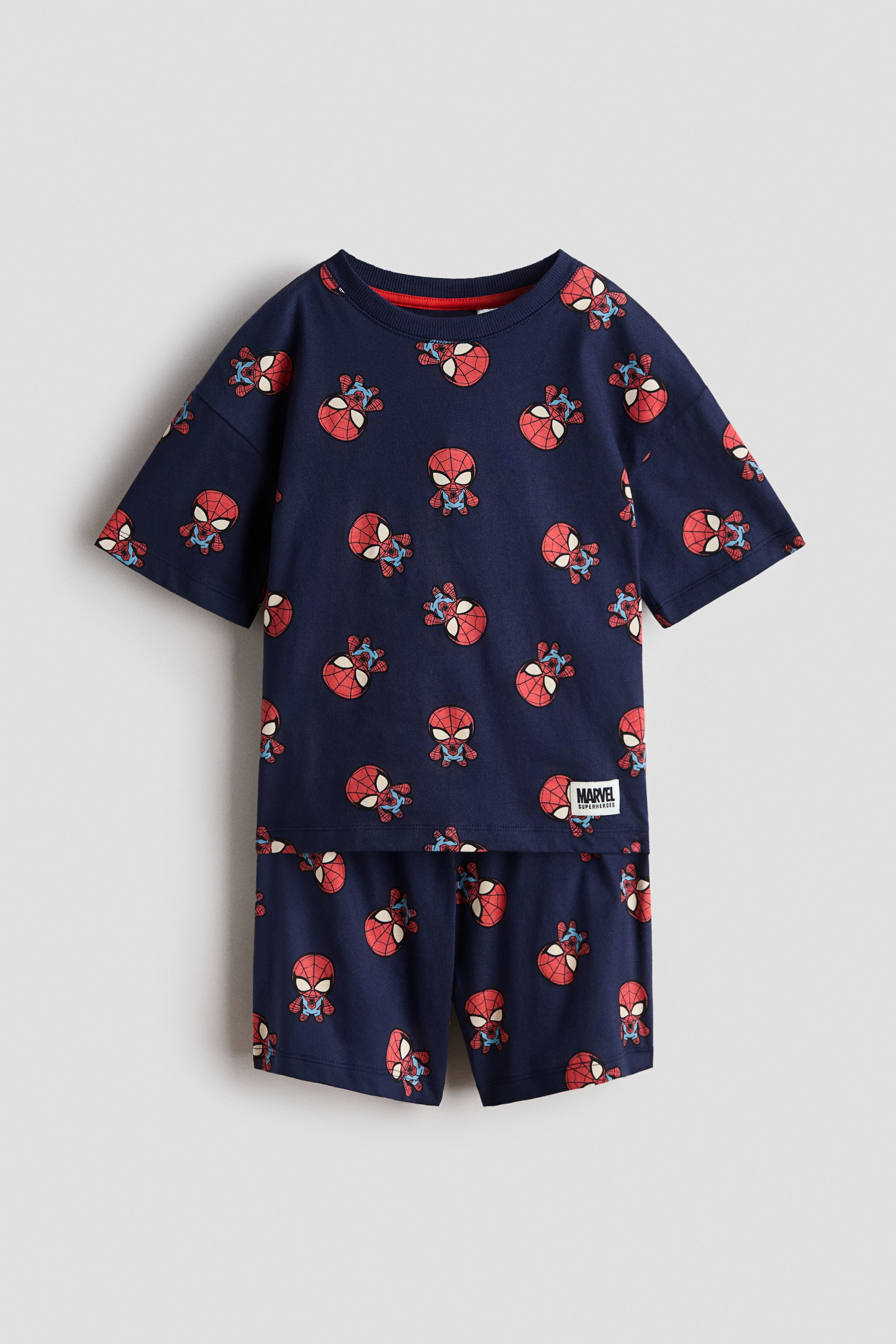 View larger image: A navy blue two-piece set, comprising a t-shirt and shorts, is presented front-facing, displaying a repeating pattern of red and blue Spider-Man faces and chibi characters. The t-shirt features short sleeves and a red ribbed neckband, with a 'Marvel Superheroes' tag on its lower right side.