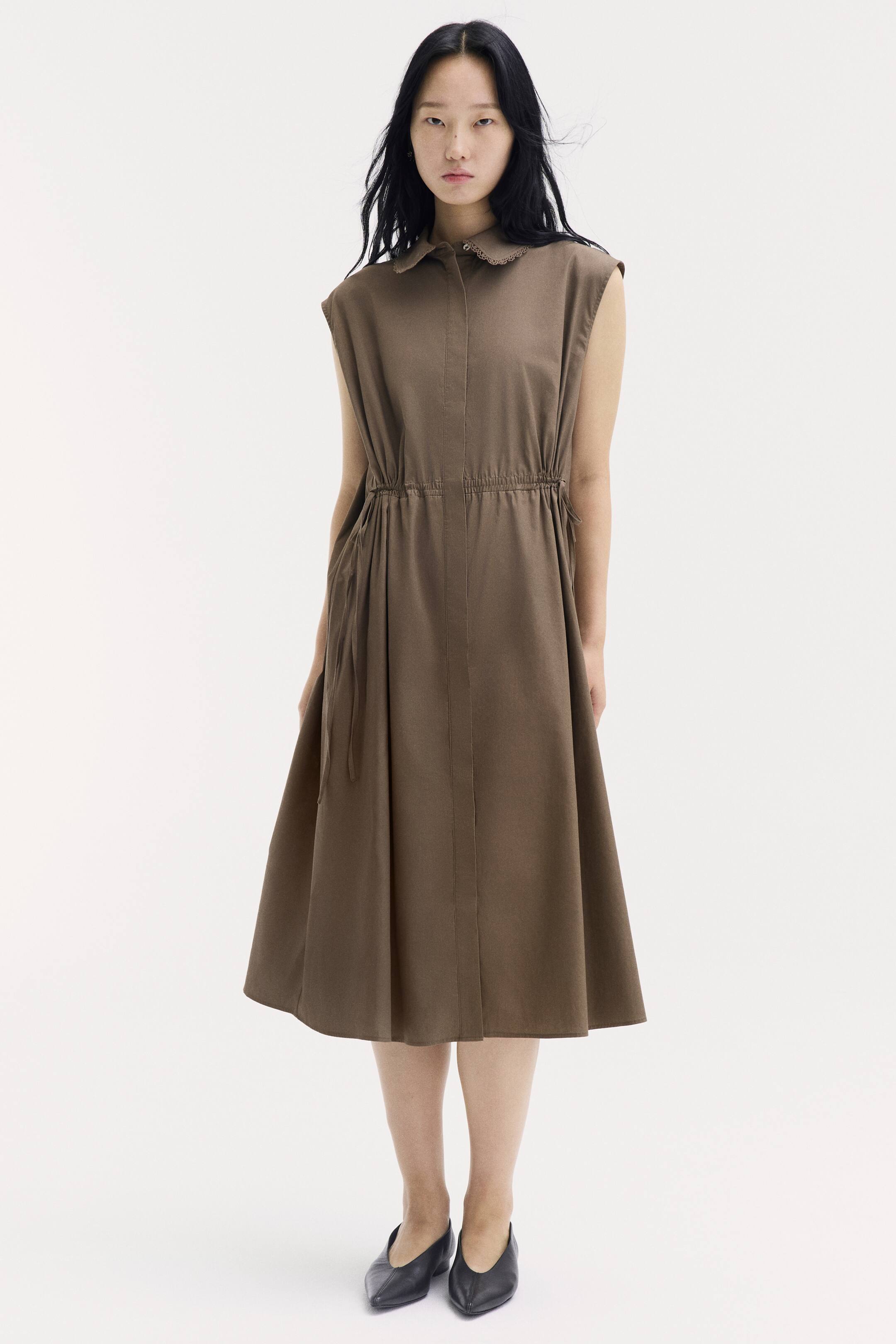 View larger image: The model is wearing a calf-length, sleeveless brown cotton dress. The dress features a lace-trimmed collar, a concealed button placket, a gathered drawstring waist, and a gently flared skirt, paired with black pointed-toe flat shoes.