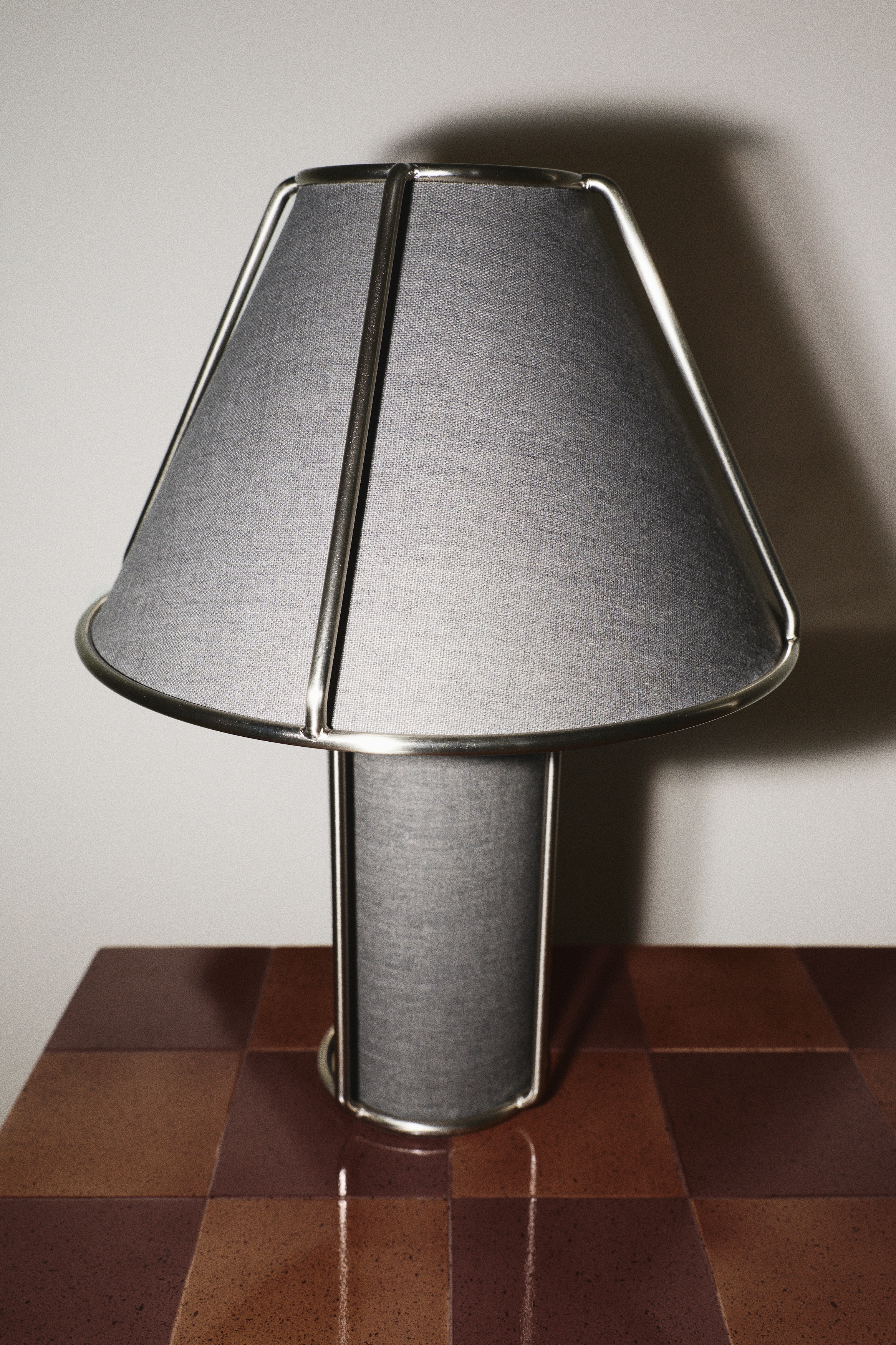 Deux lampes de table sont exposées de face, l'une plus grande et l'autre plus petite, chacune dotée d'une base cylindrique en tissu et d'un abat-jour trapézoïdal en tissu, entouré d'un cadre en fil métallique de ton argenté. La lampe plus grande présente un tissu bicolore beige clair et crème, tandis que la lampe plus petite arbore un tissu bicolore gris foncé et gris clair.