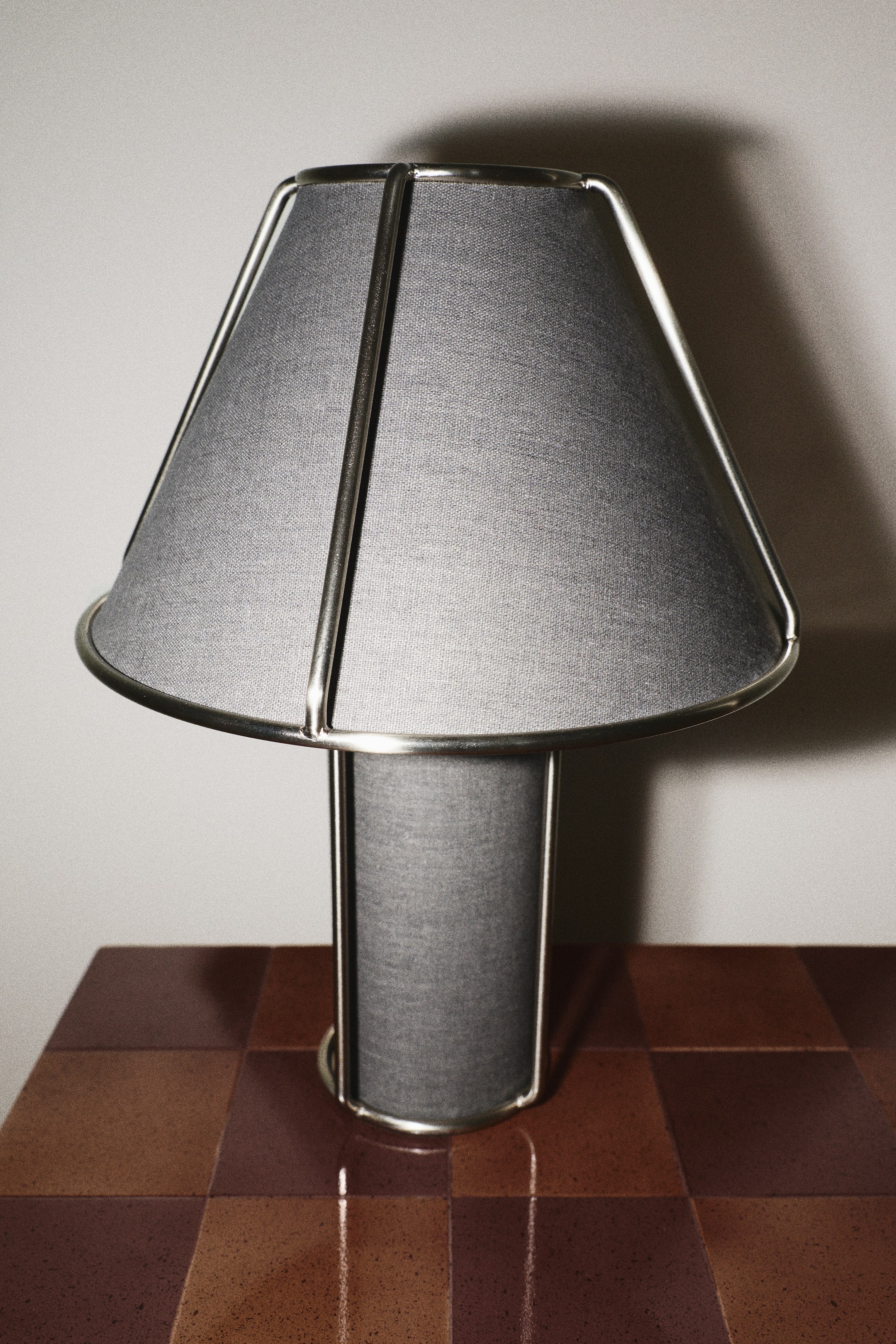 Agrandir l'image: Une lampe de table, vue de face, met en valeur un abat-jour en tissu mélange de lin gris et un pied cylindrique assorti, tous deux superposés sous une structure apparente en fil d’acier inoxydable poli.