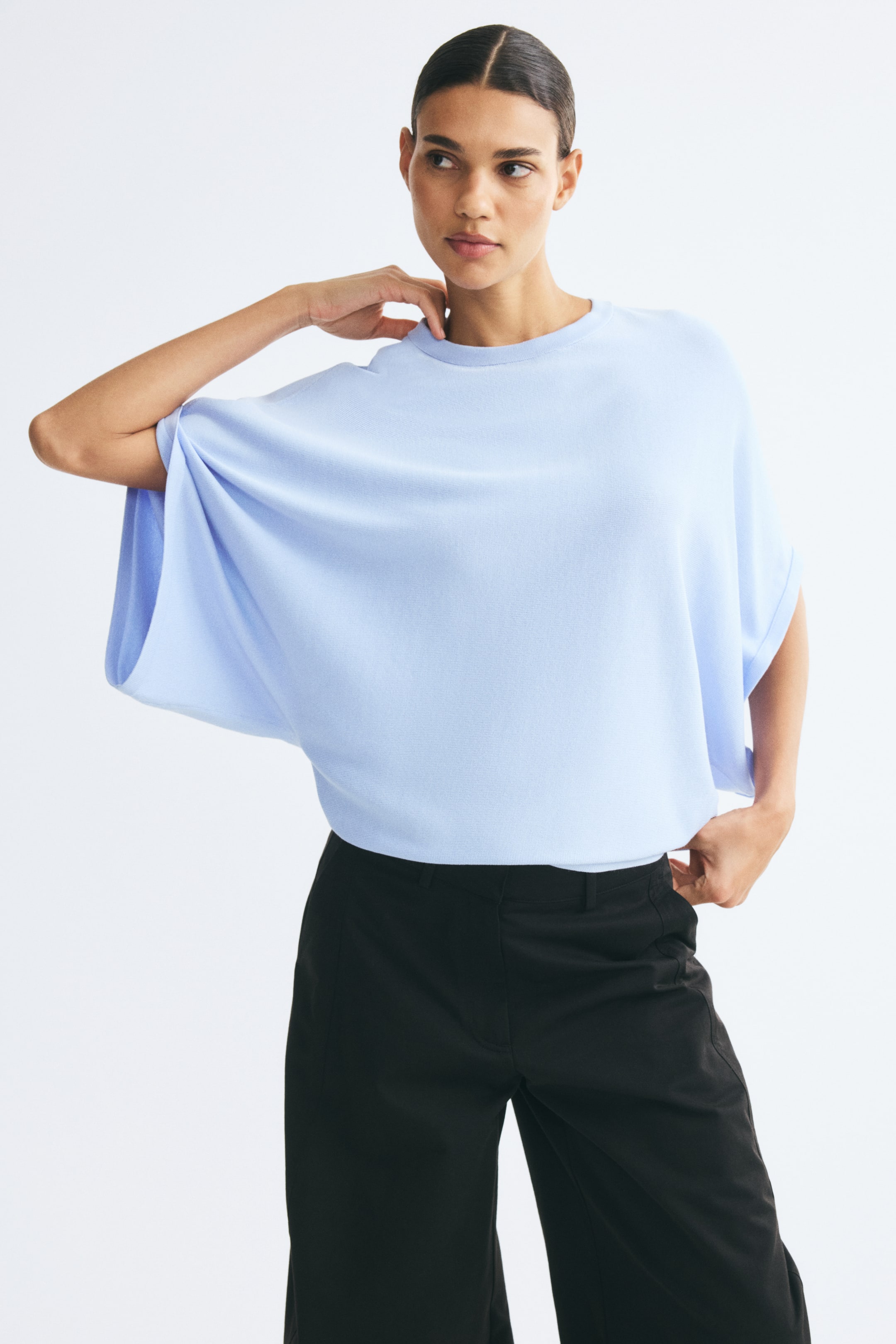 Größeres Bild ansehen: Ein hellblaues Oberteil im Boxy-Stil aus Feinstrick wird vom Model getragen und verfügt über einen Rundhalsausschnitt mit Rippstrickbesatz, extraweite kurze Ärmel und schmale Rippbündchen. Das Oberteil ist mit einer schwarzen Hose kombiniert, während das Model eine Hand auf die Schulter legt und die andere nahe dem Bund hält.