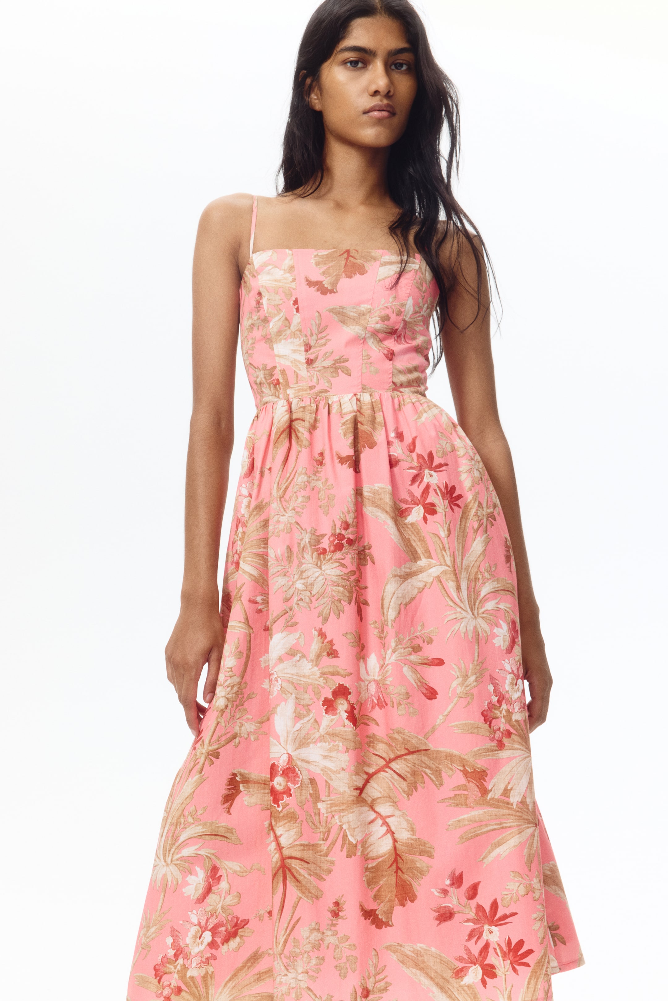 View larger image: A pink dress is displayed front-facing, featuring thin spaghetti straps, a fitted bodice, and a gathered waistline. It showcases a full A-line skirt with a tropical floral print in brown, beige, and red tones.