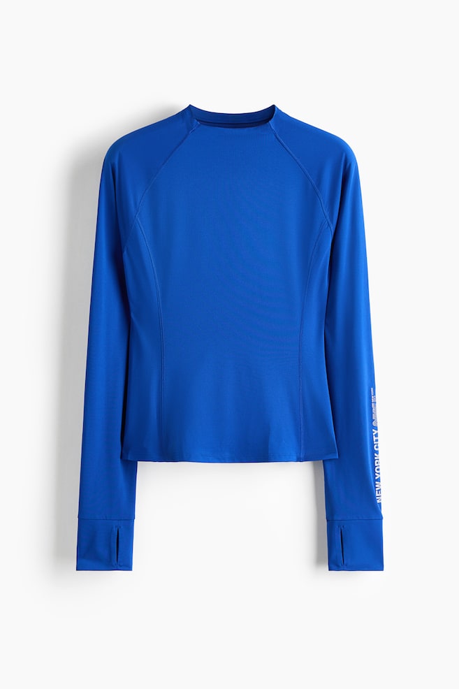 A royal blue long-sleeve athletic top is displayed from a front-facing angle, featuring a crew neck, raglan sleeves, and contoured side seams. The long sleeves are ending in wide cuffs with thumbhole details, with vertical white text printed along the lower left sleeve. - 1