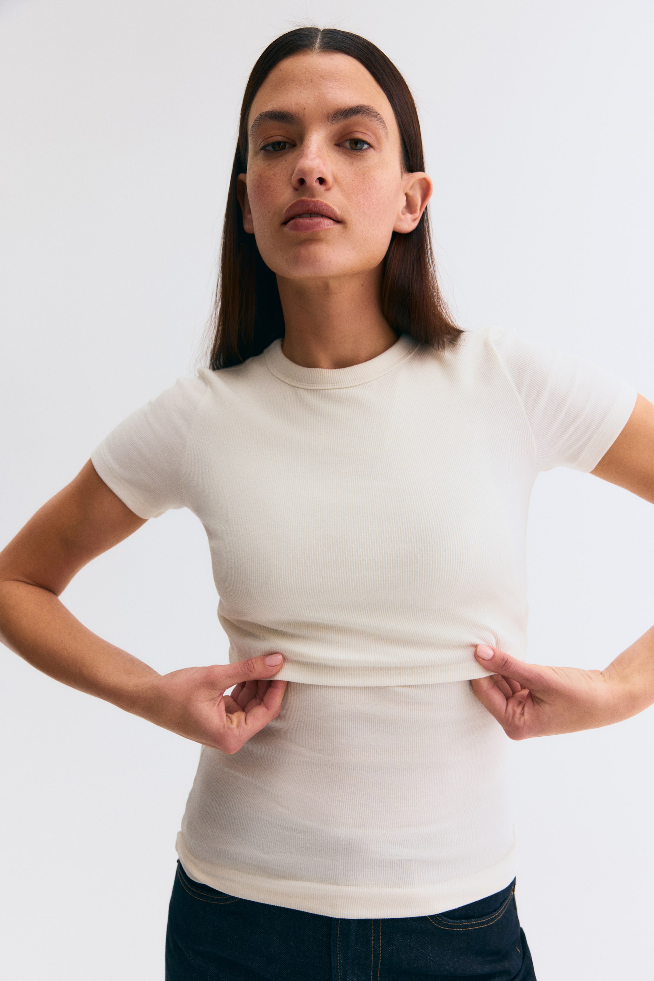 View larger image: An off-white ribbed knit, short-sleeved crew-neck T-shirt is worn, fitting close to the body. Hands gently lift the T-shirt hem, emphasising the fabric's stretch and highlighting its fine ribbed texture.