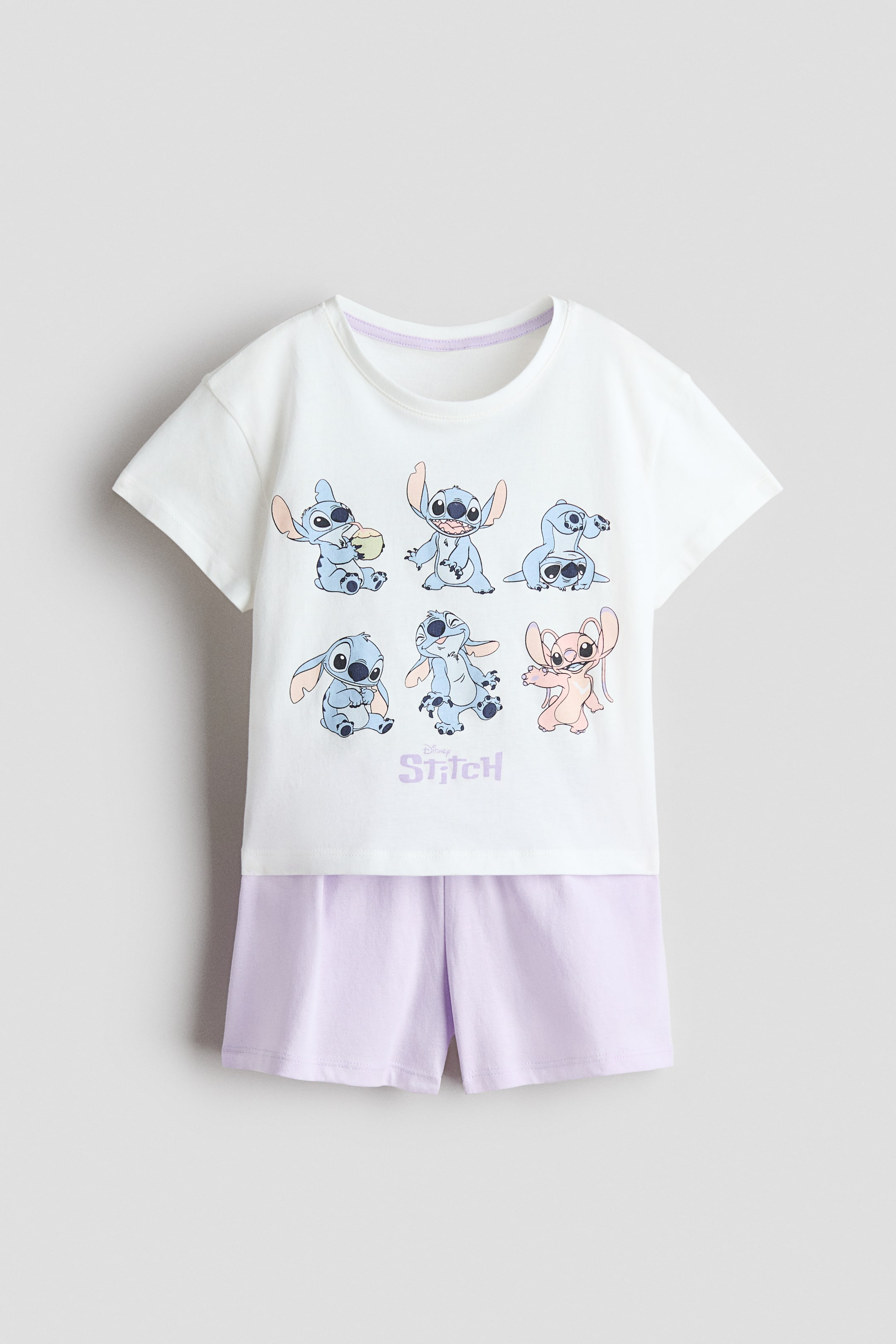View larger image: A white short-sleeved T-shirt and light-purple shorts are laid flat as a pyjama set, FRONT. The T-shirt features a graphic print of six cartoon characters, mostly blue and one in pink, with "Disney Stitch" written in purple below them. The round neckline shows a visible purple stitch along its inner rim.