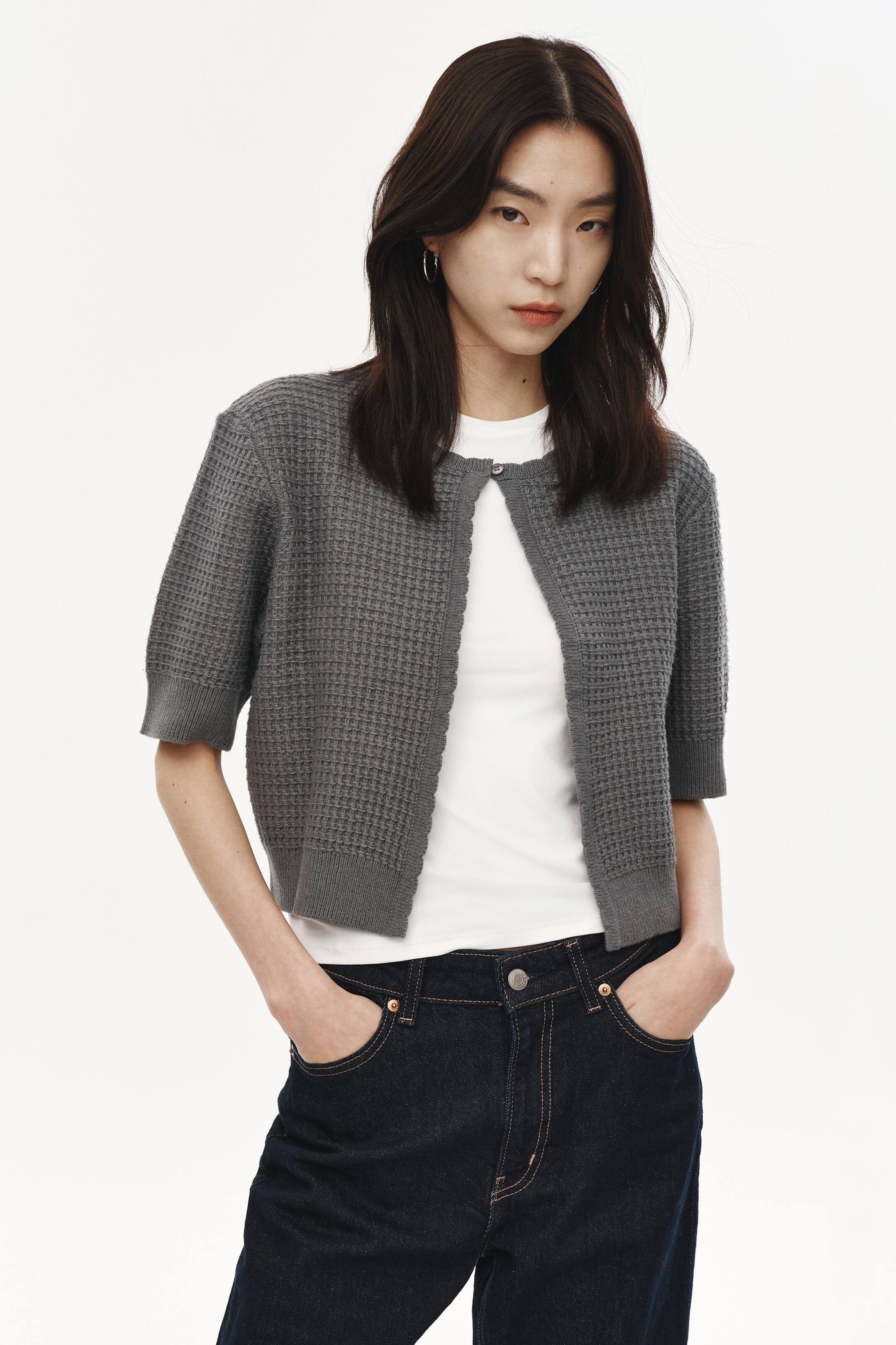 Xem ảnh lớn hơn: Một chiếc áo cardigan xám ngắn đang được giới thiệu, nổi bật với kết cấu dệt waffle và tay ngắn có viền bo. Áo được mặc mở bên ngoài áo thun trắng, có một nút cài duy nhất ở trên cùng và gấu áo bo viền.