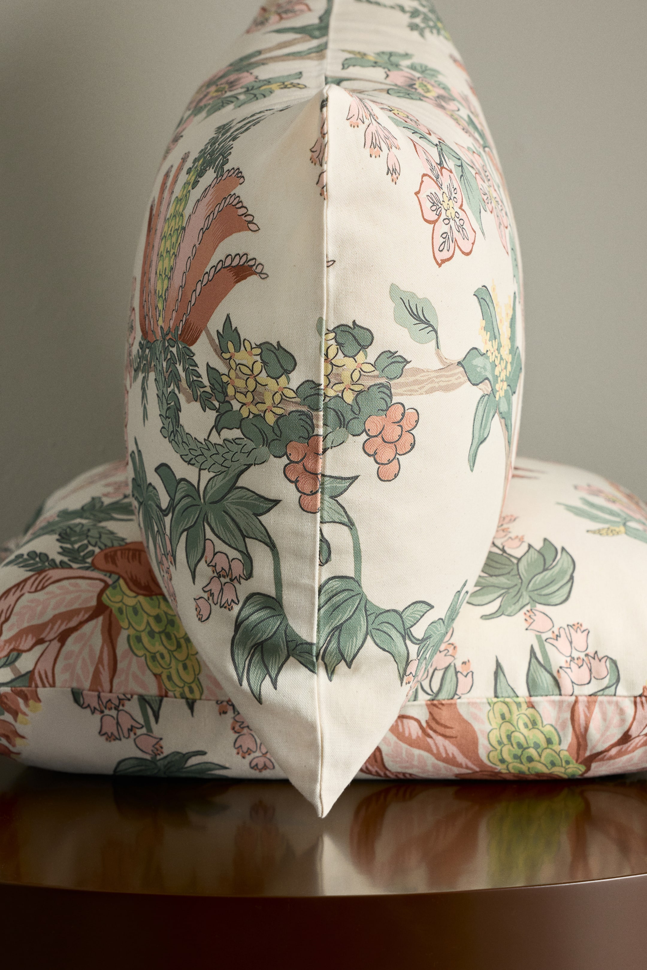 View larger image: Two decorative cushions feature a detailed floral pattern. One cushion stands upright, angled slightly to the right to show its seamed edge, while the other lies flat behind it. The off-white fabric is printed with large green leaves, pink and yellow blossoms, and clusters of orange berries.