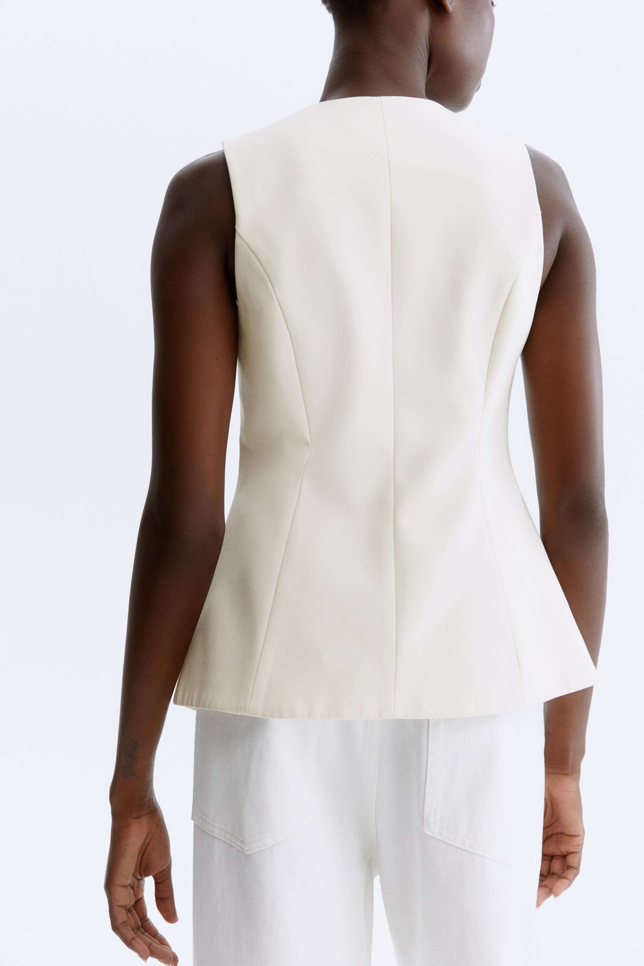 Ampliar la imagen: Un top sin mangas de color blanco roto se ve desde la parte posterior, con una silueta ajustada alrededor de la cintura que se ensancha en un dobladillo peplum. Las costuras verticales de forma corren a lo largo de la espalda, y se lleva sobre pantalones blancos.