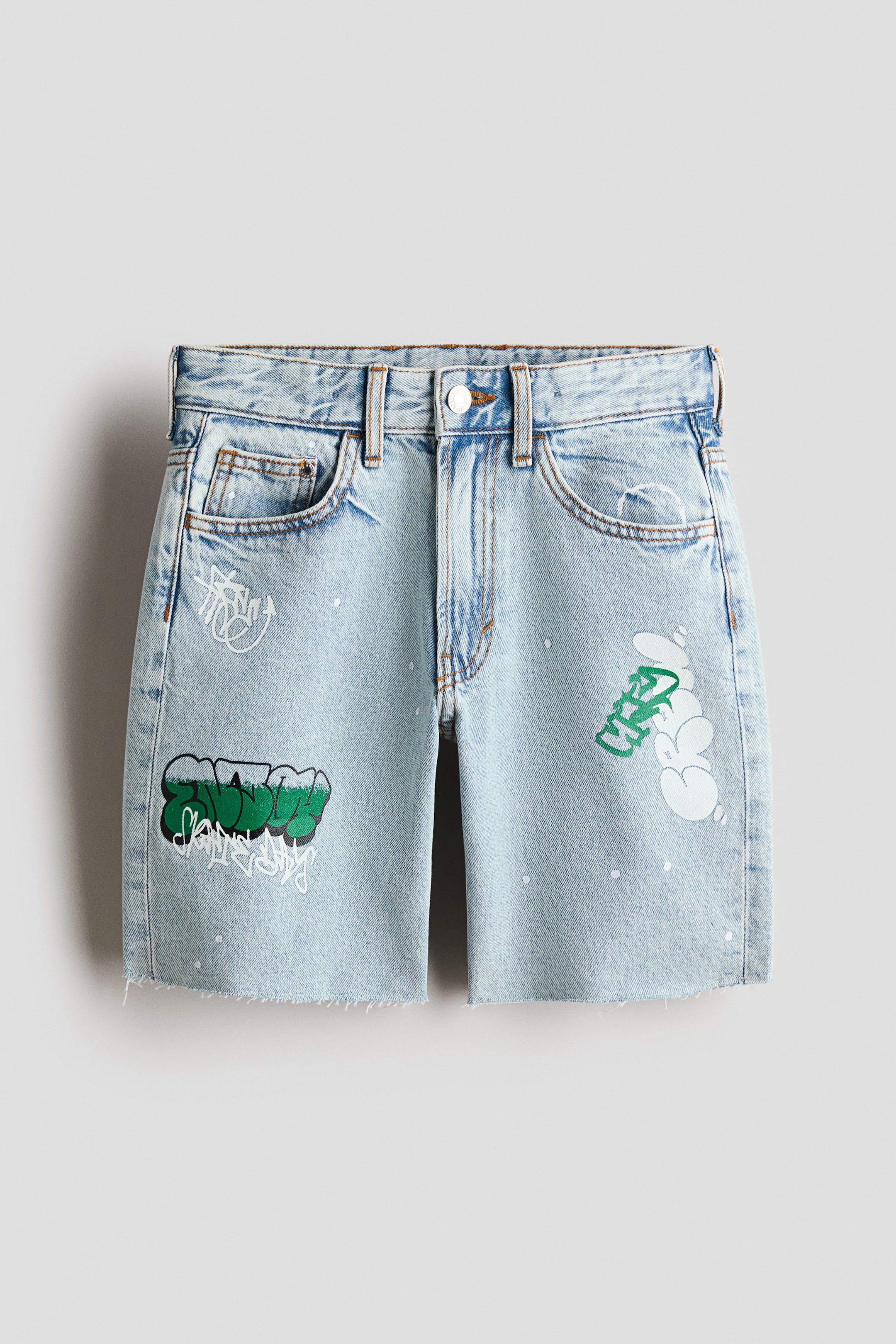 Vezi imaginea mai mare: Pantaloni scurți cu 5 buzunare, orientați spre față, din denim moale, albastru deschis, prezintă o croială lejeră în zona șoldurilor și coapselor, o talie elastică ajustabilă cu fermoar și închidere cu nasture, și un tiv nefinisat. Imprimeuri în stil graffiti în verde, negru și alb detaliază partea din față, alături de mici stropi de vopsea albă și zone uzate.
