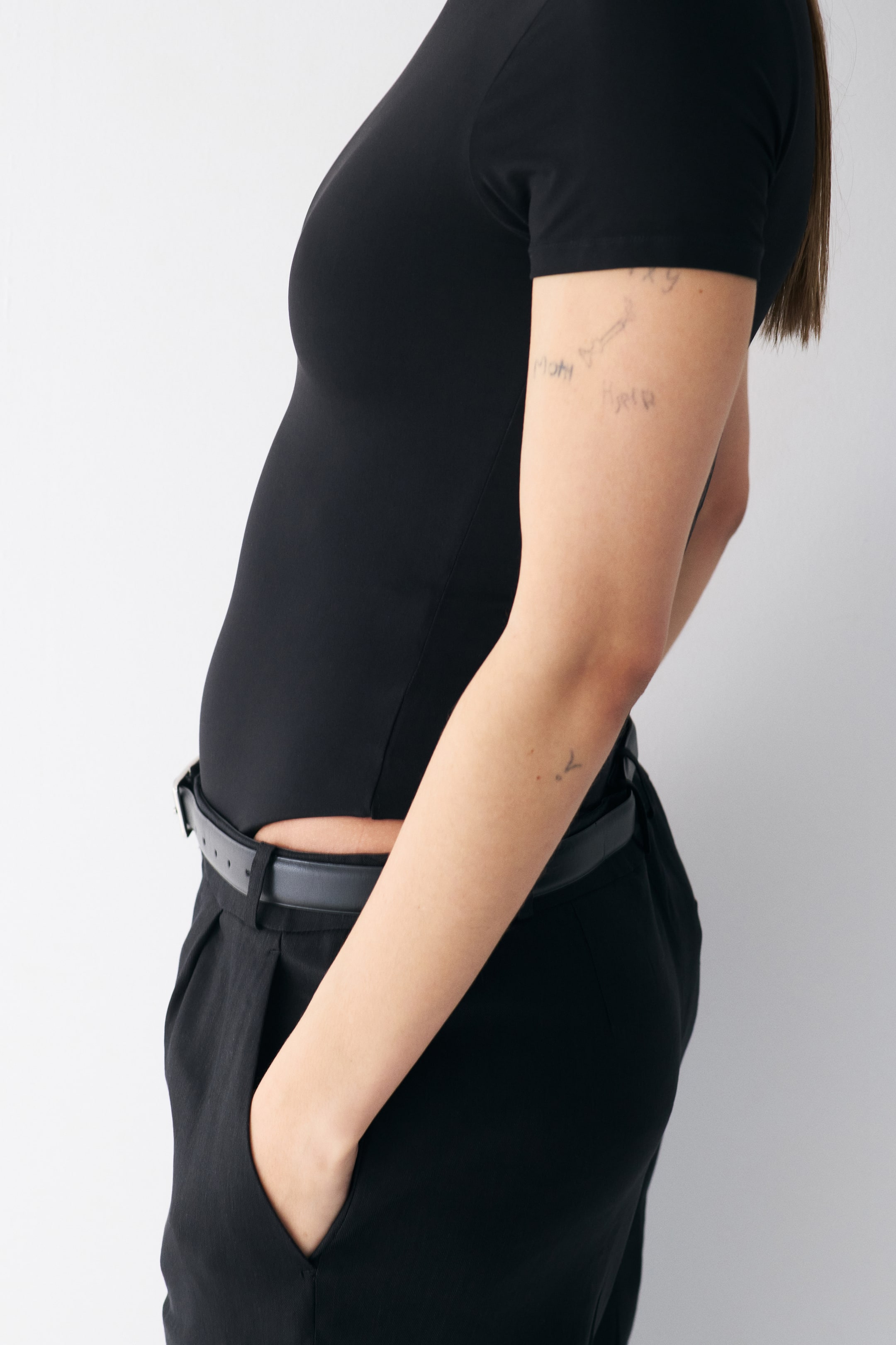 Visualizza immagine più grande: Un top nero a maniche corte aderente al torso, visibile da una vista laterale. È indossato infilato in pantaloni neri a pieghe, fissati con una cintura nera intorno alla vita.