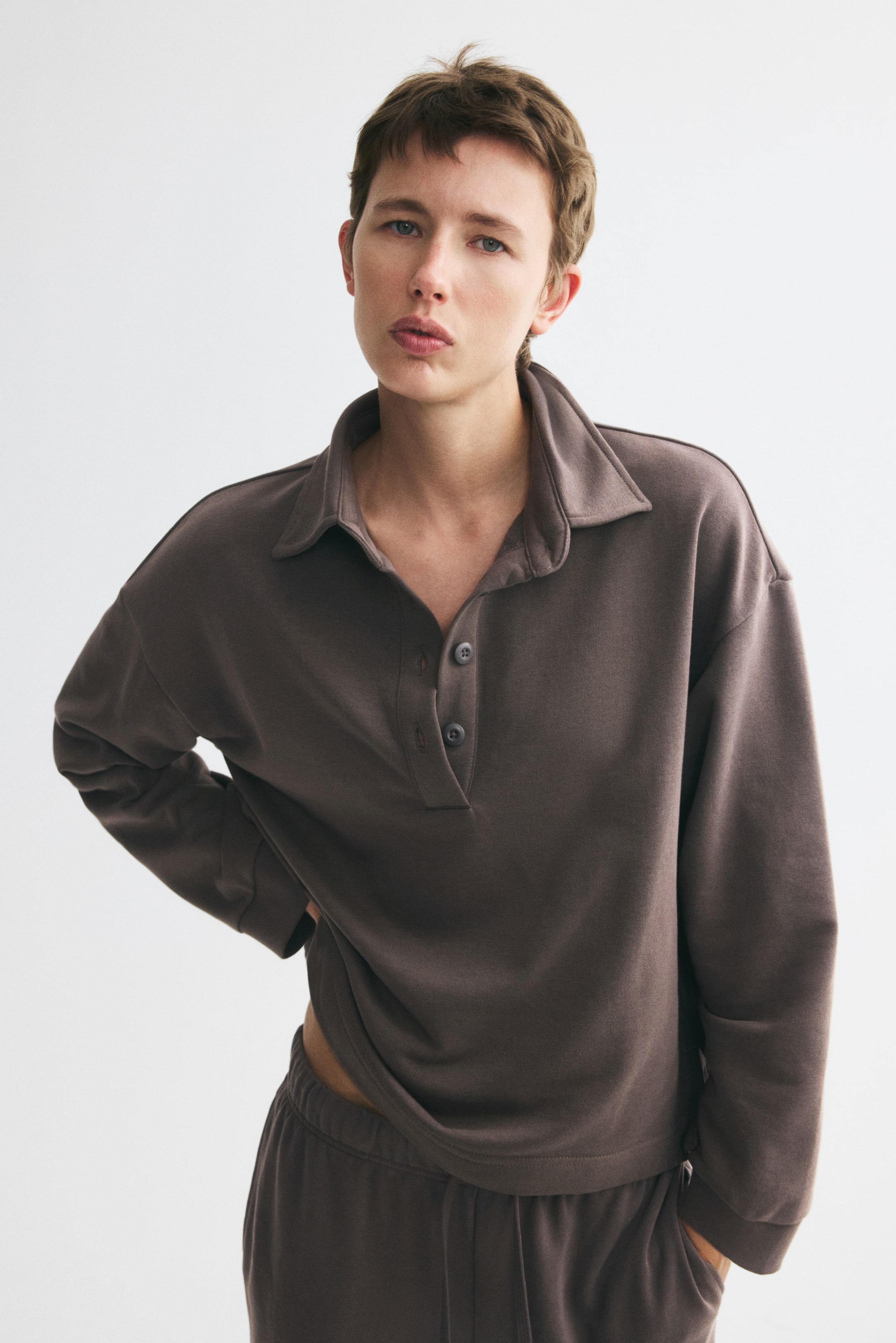 View larger image: The model is wearing a top in lightweight sweatshirt fabric, featuring a collar, a three-button placket, dropped shoulders, and long sleeves with ribbed cuffs. The model is slightly adjusting the top's straight-cut hem, which features side slits, and is also wearing matching sweatpants.