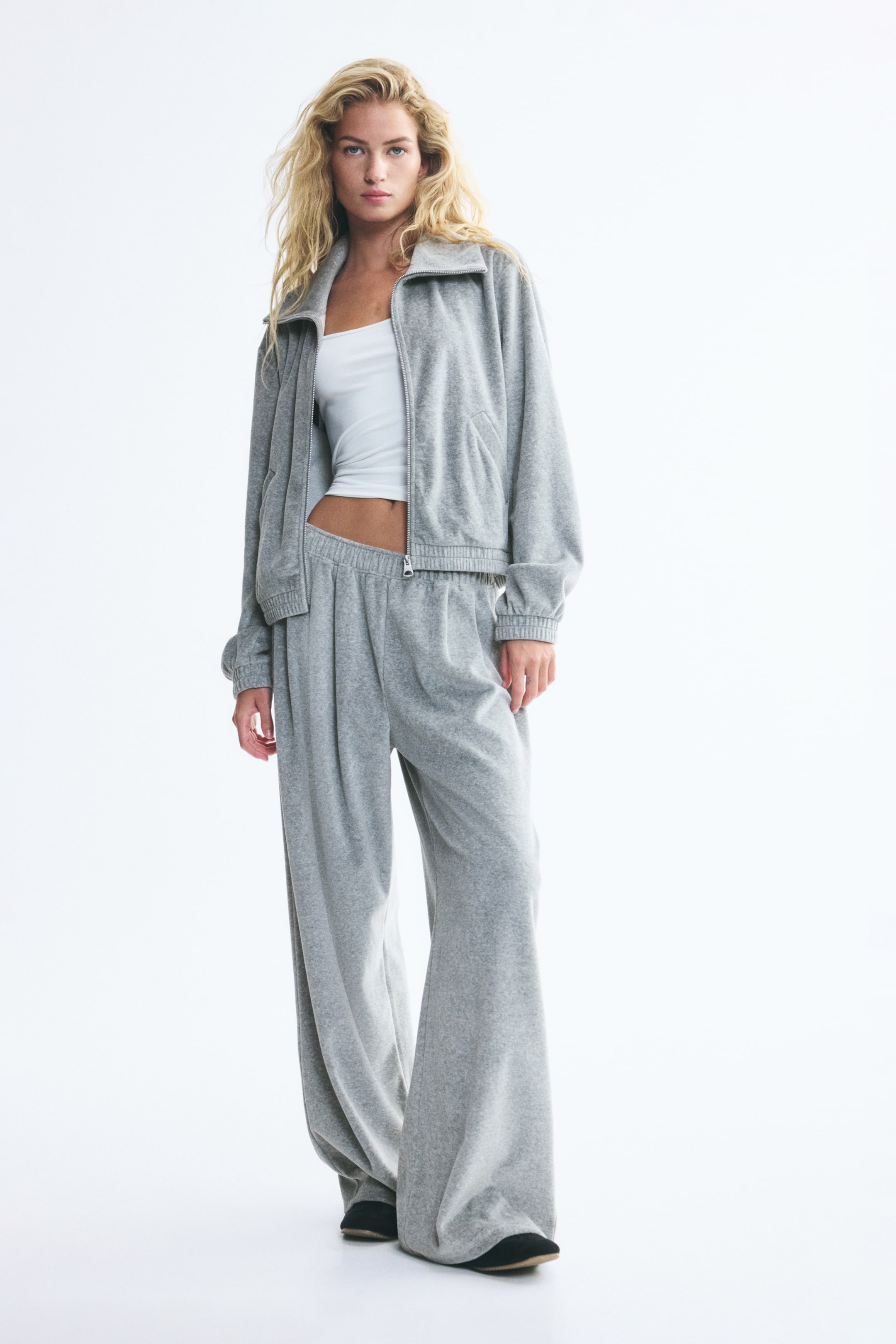 View larger image: The model is wearing grey cotton-blend velour joggers featuring an elasticized waistband and straight legs with front pleats. The joggers are paired with a matching grey velour zip-up jacket and a white cropped tank top.