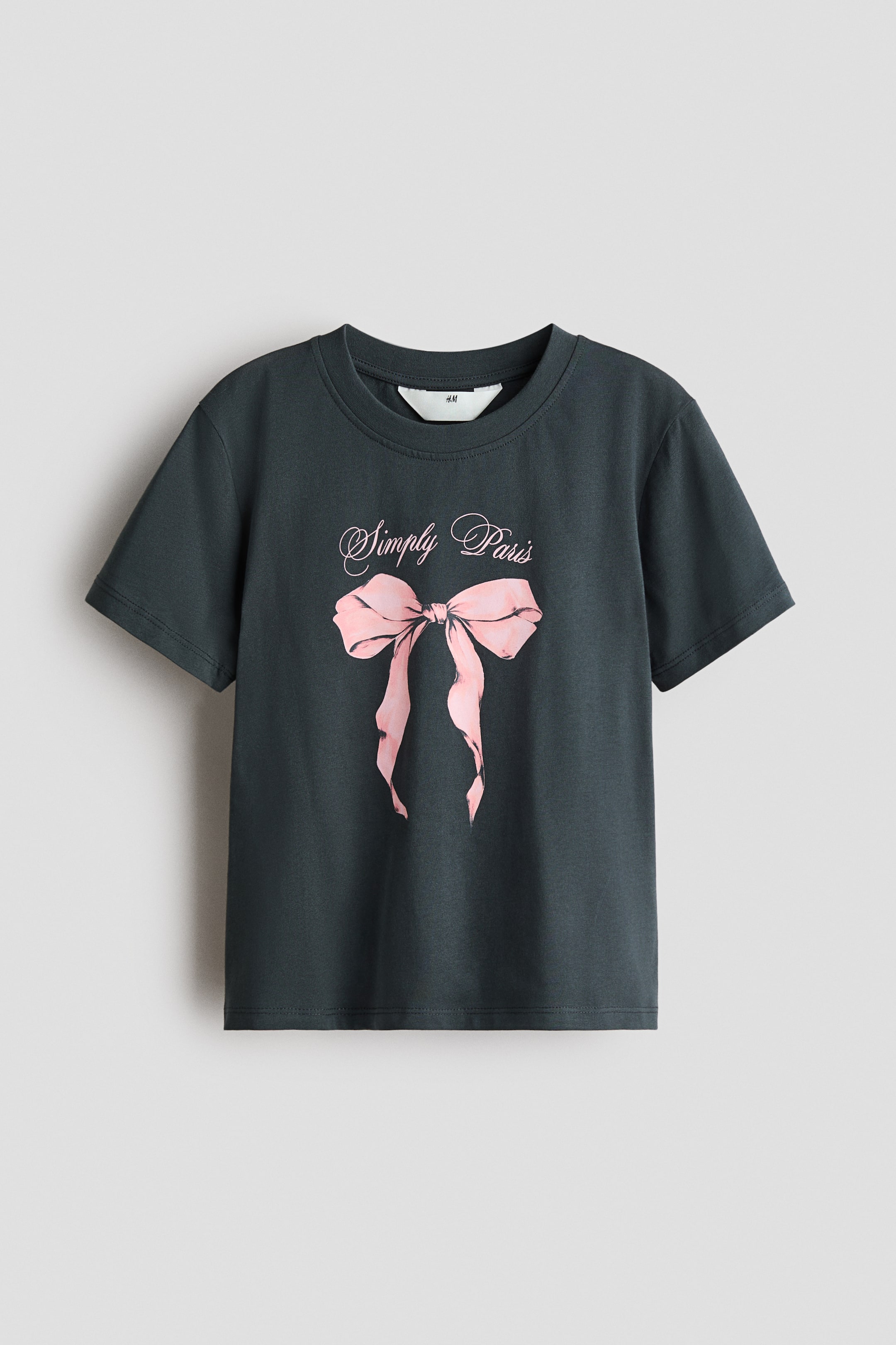 Visa större bild: En kolgrå T-shirt visas framifrån, med ribbad kant runt halsringningen och korta ärmar. Ett rosa grafiskt tryck, som föreställer en rosett med den kursiva texten 'Simply Paris' ovanför, syns på framsidan.