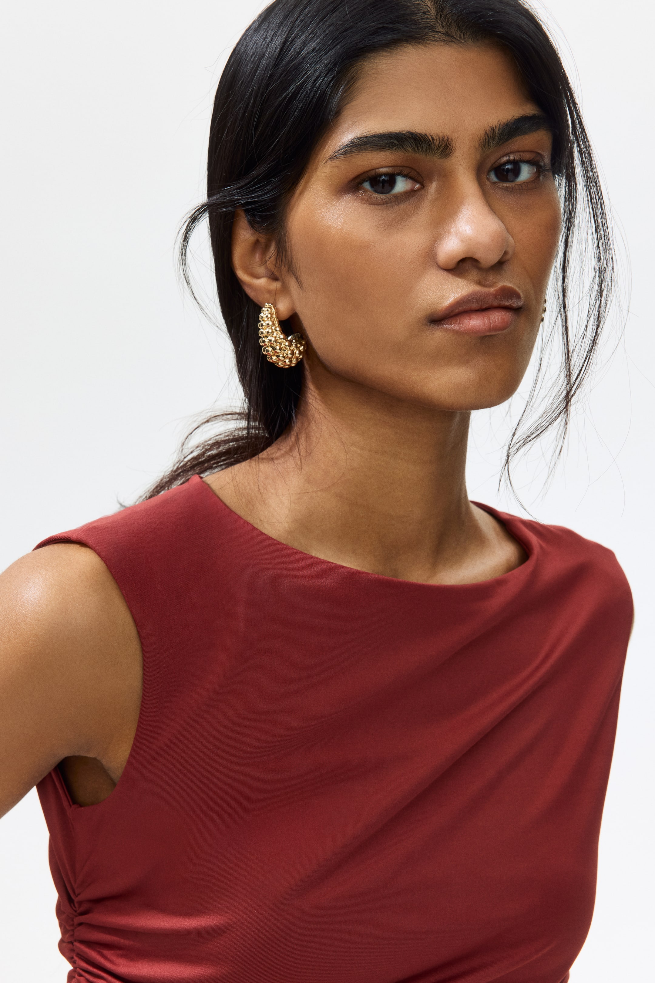 View larger image: The model is wearing a snug, sleeveless top in a rich maroon jersey with a subtle sheen. It boasts a boat neckline and a ruched seam on one side of the waist, delivering a softly draped look.