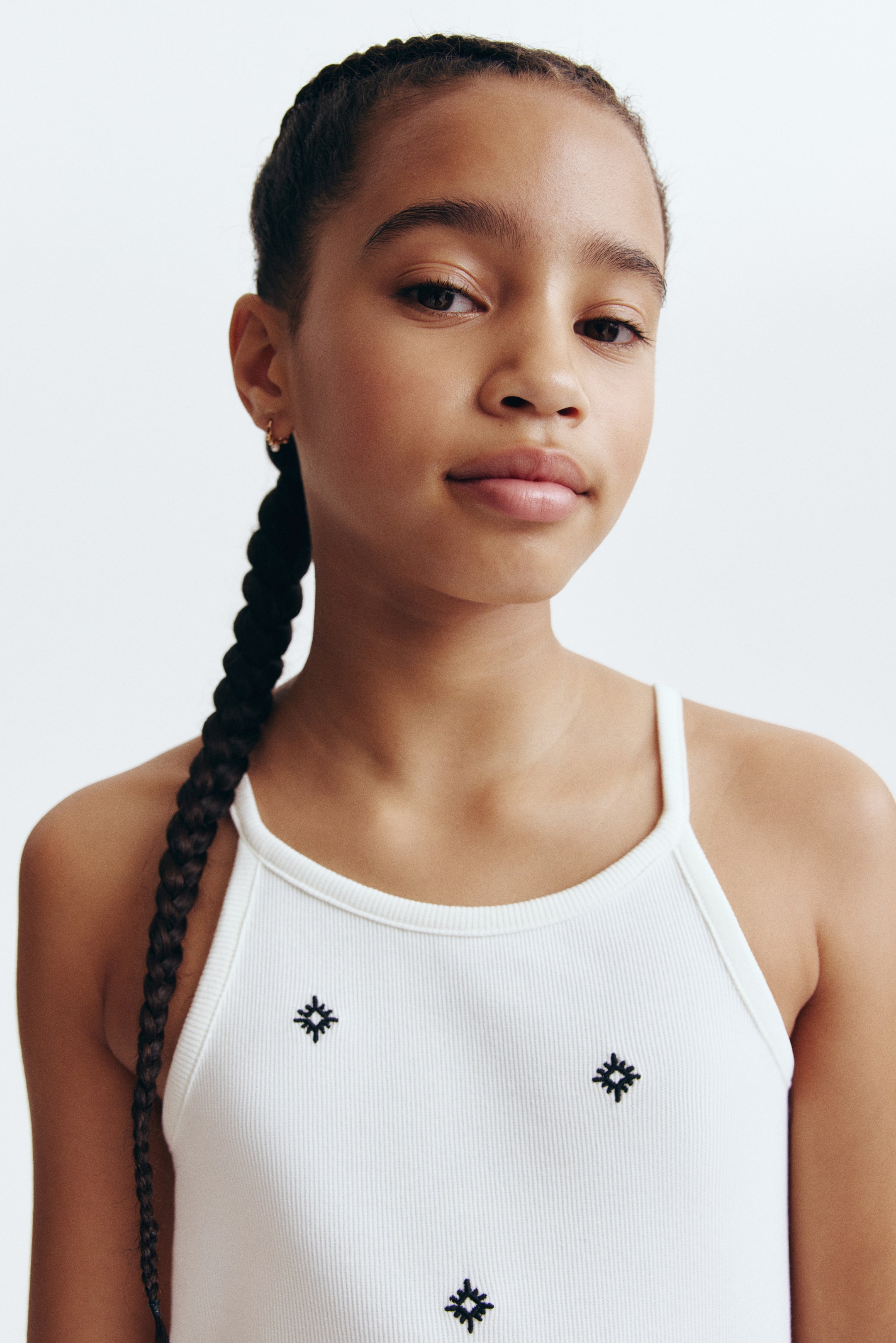 View larger image: A white ribbed knit vest is being presented, featuring thin shoulder straps and multiple small, dark embroidered geometric patterns on its front.