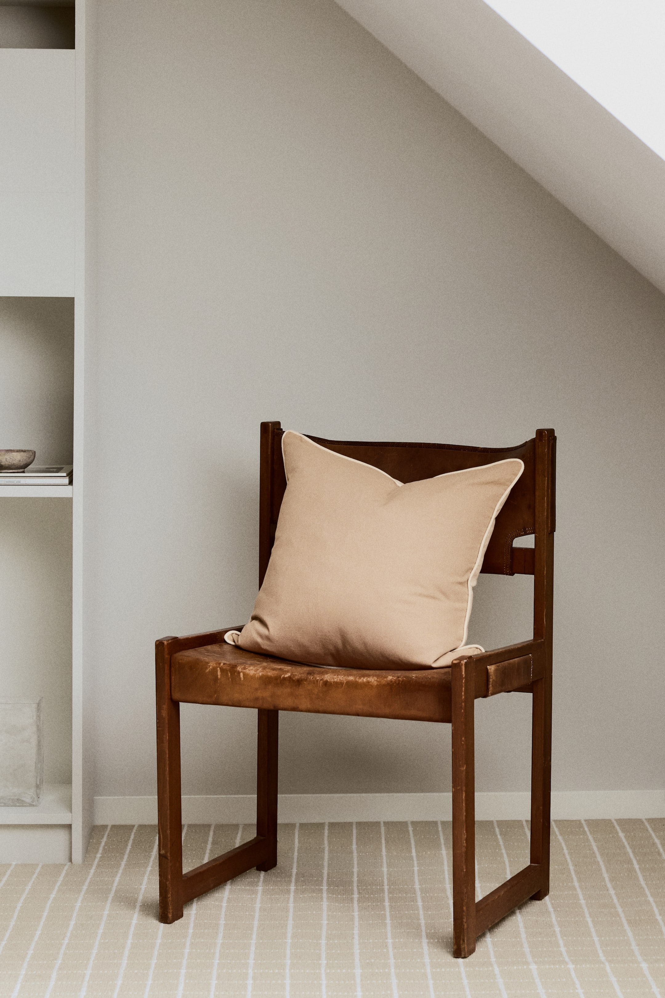 Voir une image plus grande: Un coussin carré de couleur beige clair est présenté à la verticale, avec un passepoil crème le long de tous ses bords. Sa surface avant est légèrement orientée vers la gauche, affichant une texture de tissu uni.