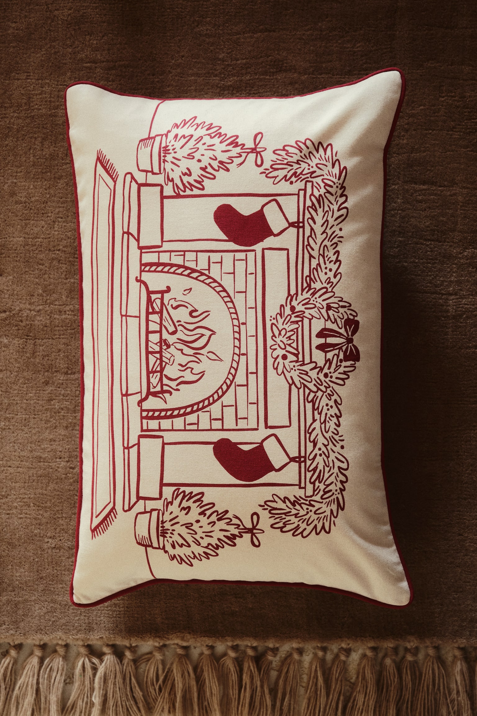 A rectangular off-white lumbar pillow is shown front-facing, showcasing a red line-drawn illustration of a cosy fireplace scene. The illustration features a burning fireplace adorned with a mantel garland, a central wreath, hanging stockings, and potted evergreen trees on either side, all framed by red piped trim along the edges. - 1