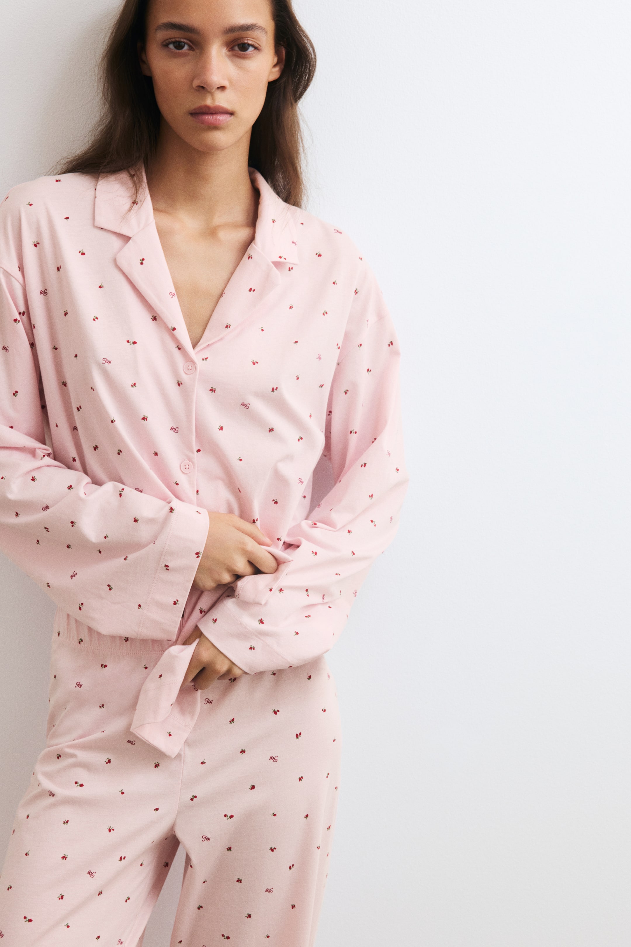 View larger image: A light pink pyjama shirt and pants set featuring a small red print is worn by the model. The long-sleeved shirt features a resort collar, dropped shoulders, and a button-down front, which the model is holding at the waist, while the matching pants feature an elasticated waistband.