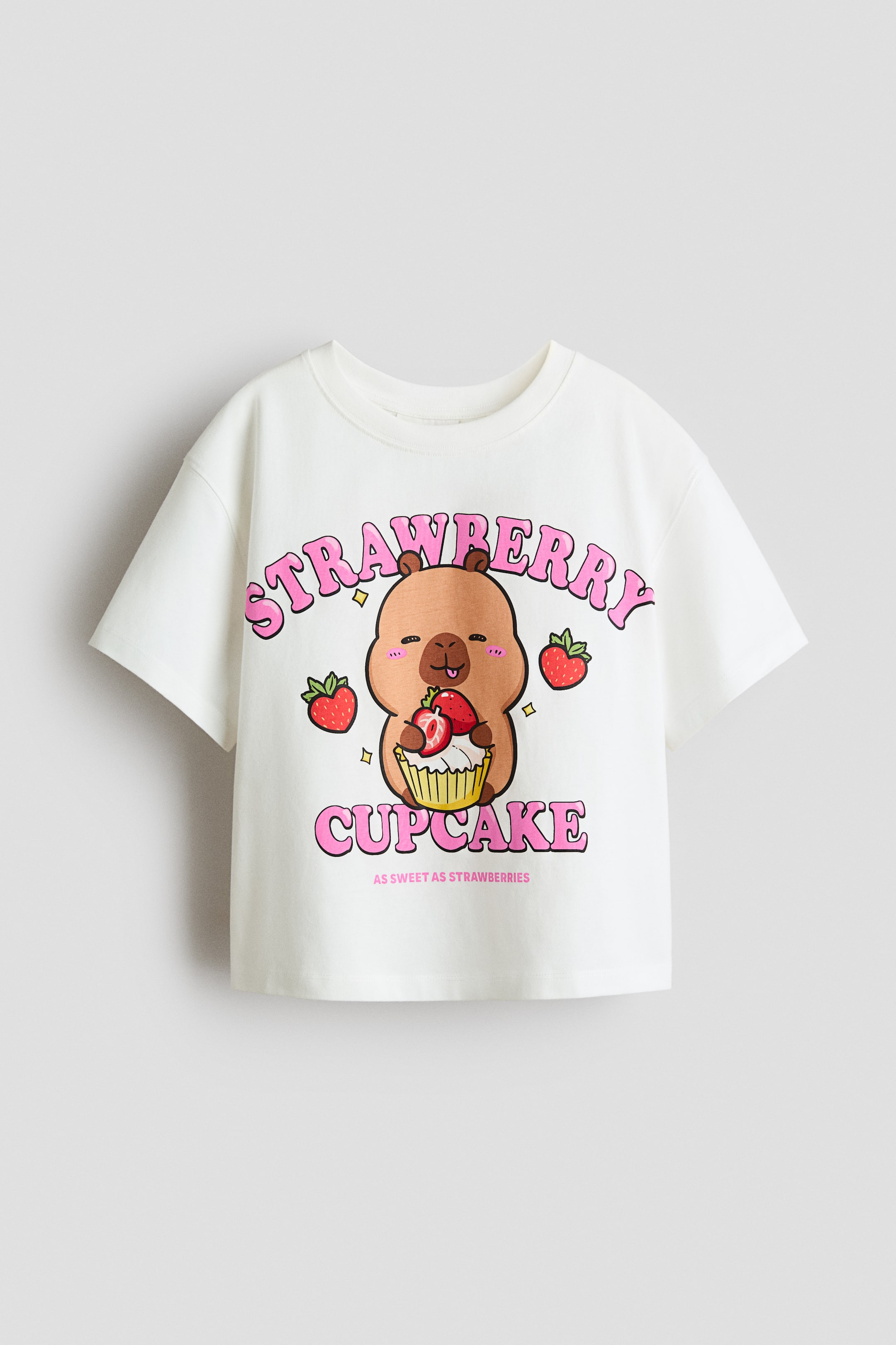 Visa större bild: En vit bomullsjersey topp visar sin framsida, med ett färgglatt tryckmotiv av en brun tecknad figur som håller en jordgubbsmuffin, med orden "strawberry cupcake" ovanför och nedanför. Den är designad med en rund, ribbkantad halsringning, nedhasade axlar och korta ärmar.