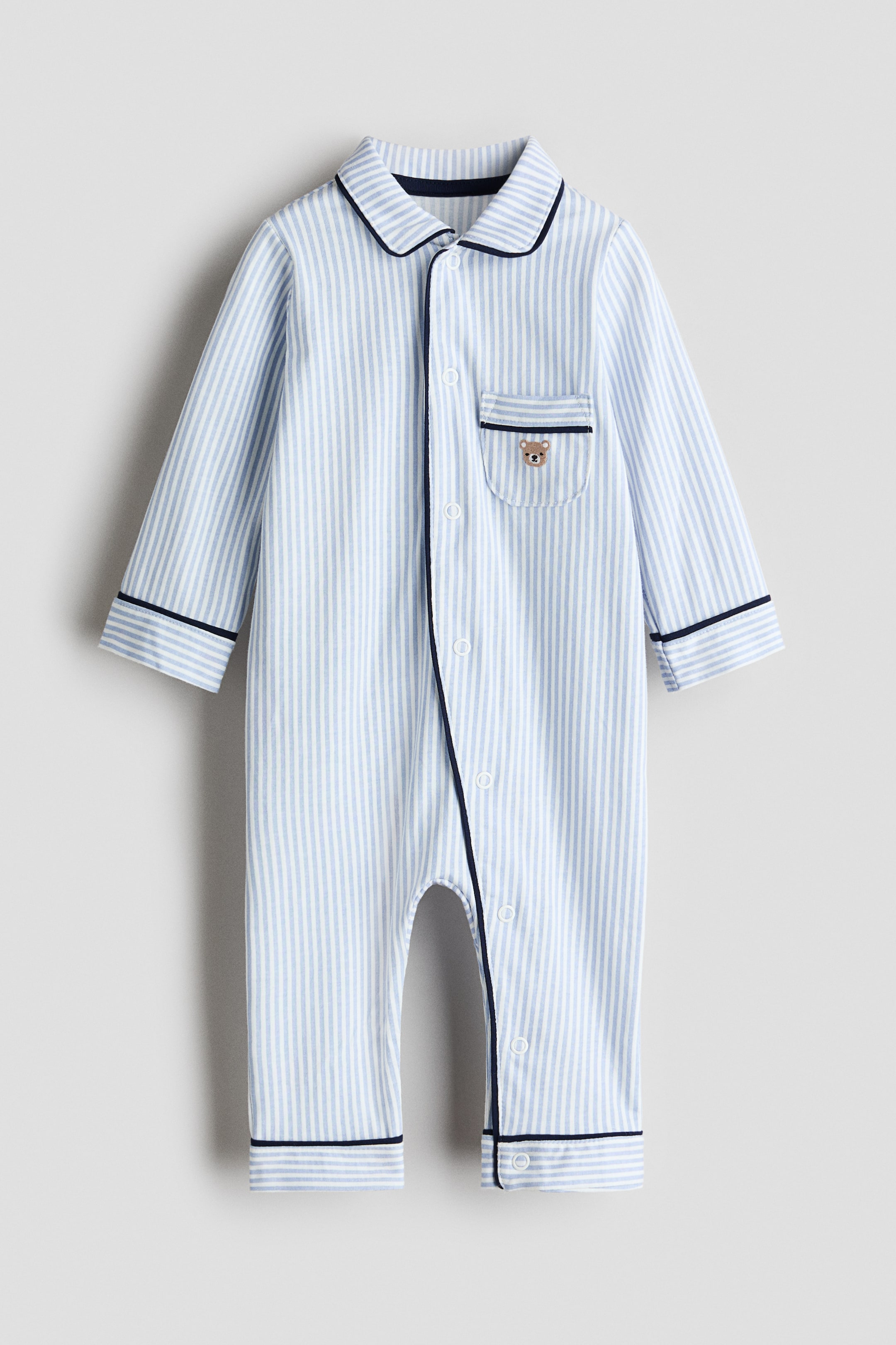 Grotere afbeelding bekijken: Een lichtblauw-wit gestreept katoenen sleepsuit wordt van voren getoond, met een ronde kraag, een klein borstzakje met een geborduurde teddybeer, en een drukknoopsluiting die aan de voorkant en langs één pijp loopt. Donkerblauwe biesjes accentueren de kraag, manchetten, het zakje, de middenbies en de broekspijpen.