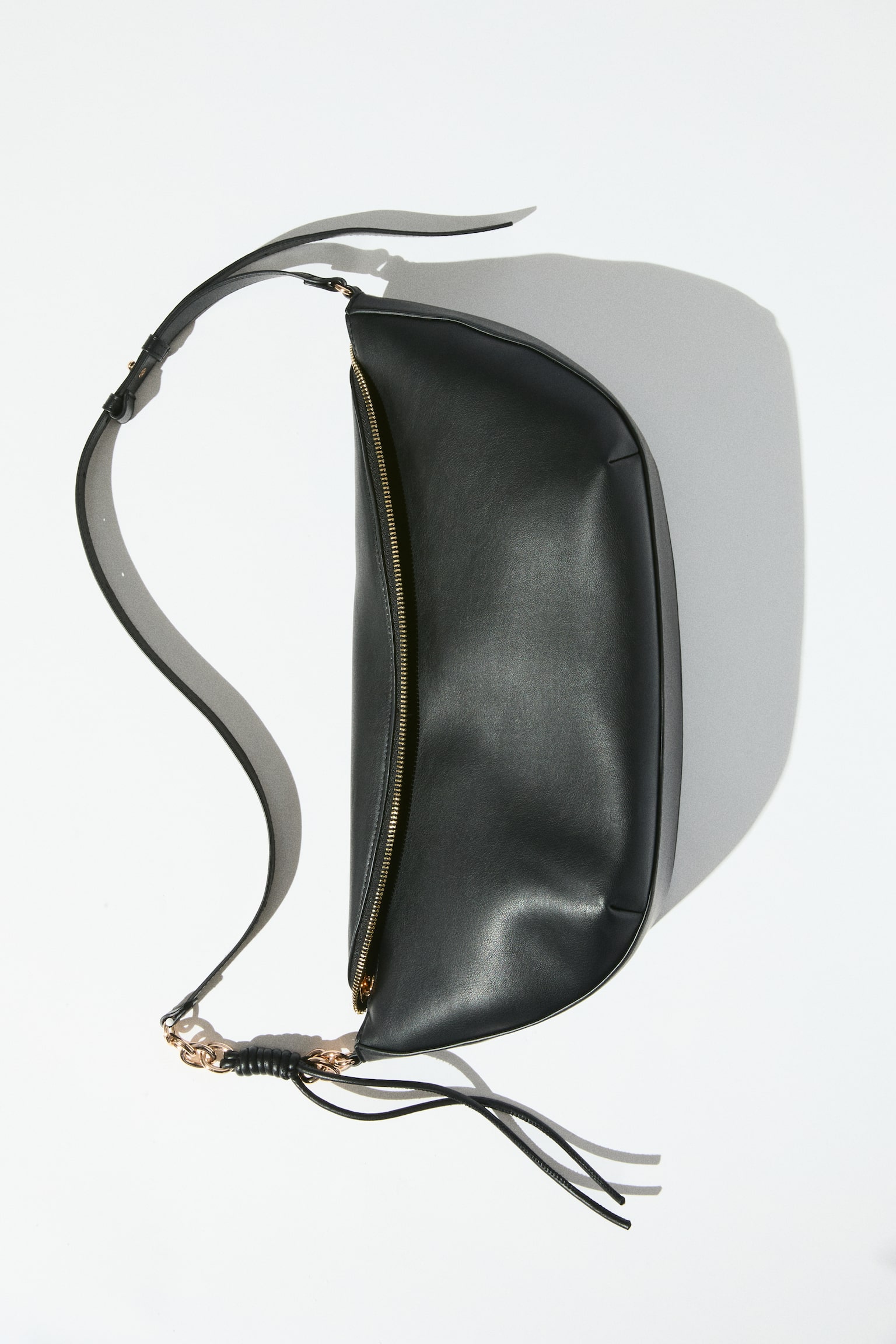 The black, crescent-shaped handbag features a smooth, supple material and a gold-toned zip closure along its top. An adjustable shoulder strap connects to the bag with gold-toned chain links and a wrapped detail on one side, as the bag is presented from a slightly elevated front angle. - 1