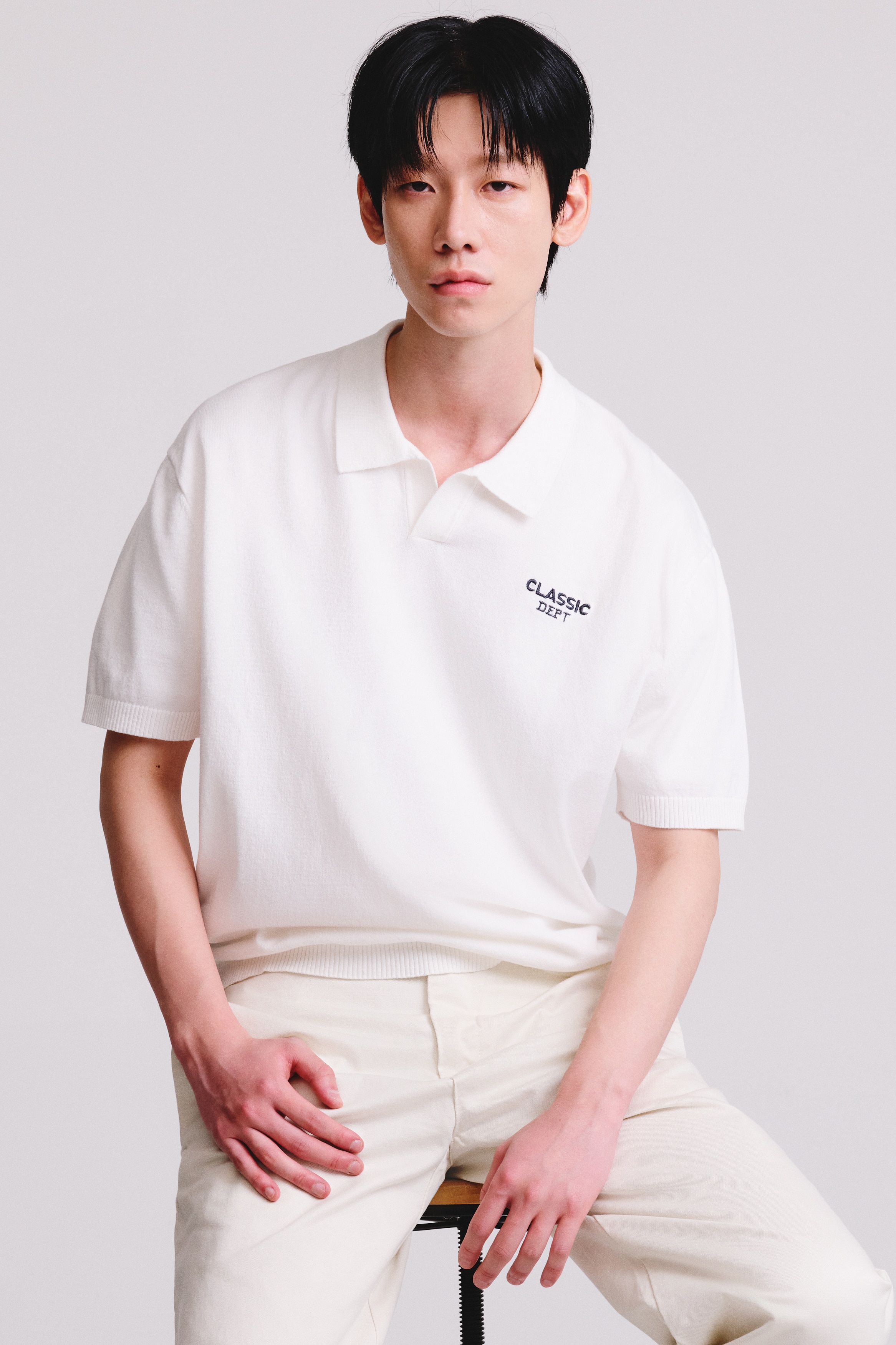 An off-white, fine-knit polo shirt is shown from the front, featuring an open collar, short sleeves, and ribbed cuffs and hem. The shirt displays the black embroidered text "CLASSIC DEPT" on the left chest.