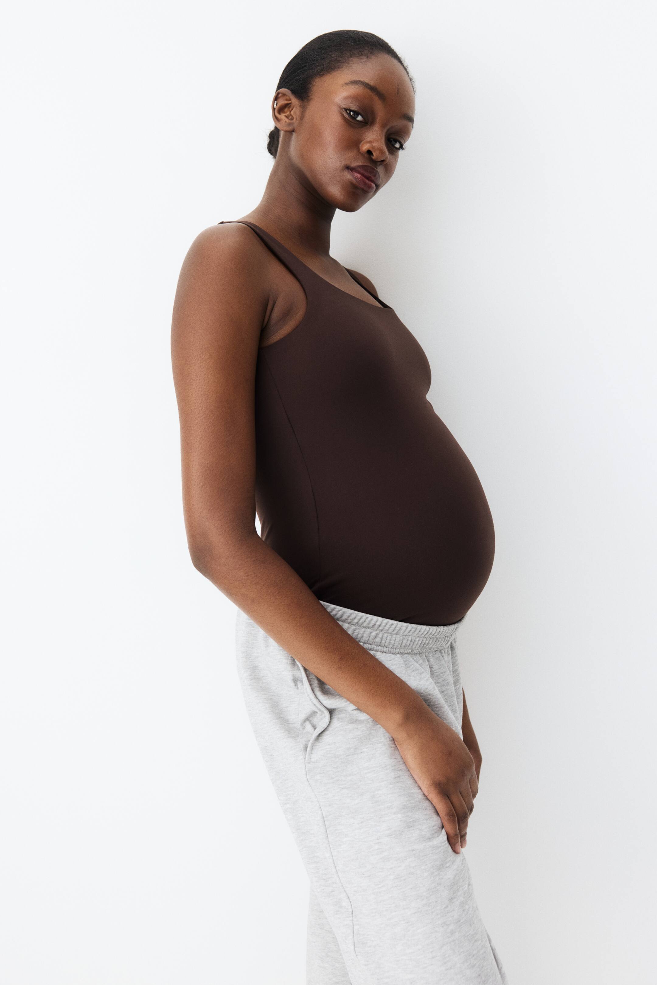 Ampliar la imagen: Una blusa de maternidad sin mangas de color marrón oscuro con tirantes finos se ajusta suavemente sobre un vientre embarazado, mientras que unos pantalones de chándal gris claro con una cintura elástica acanalada y bolsillos laterales se usan abajo. Ambas prendas están ligeramente inclinadas hacia la derecha.