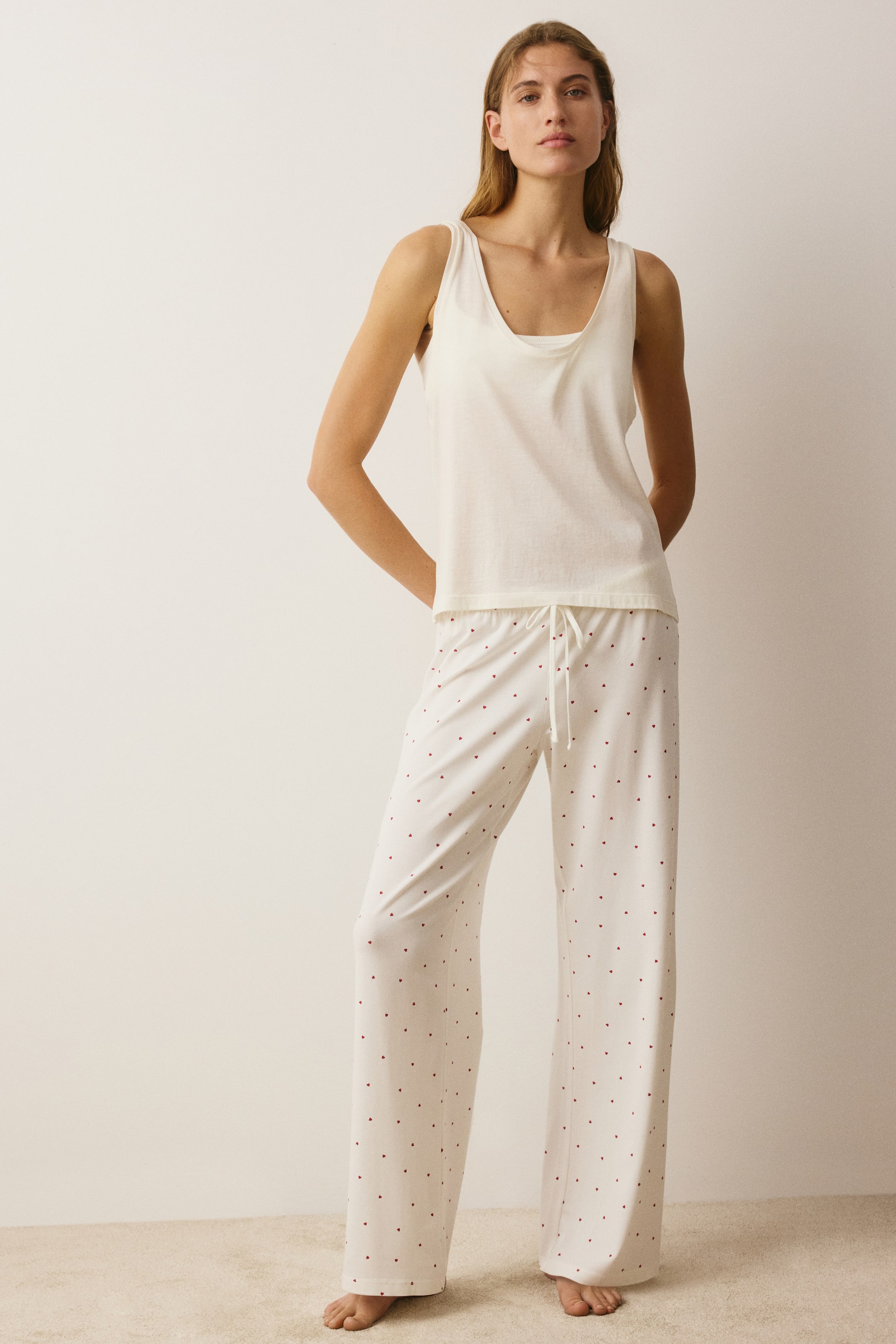 View larger image: A model is wearing loose-fit pyjama trousers in cream-coloured jersey with a small red heart pattern, facing front to show the elasticated waist with a white drawstring and the wide-leg drape.