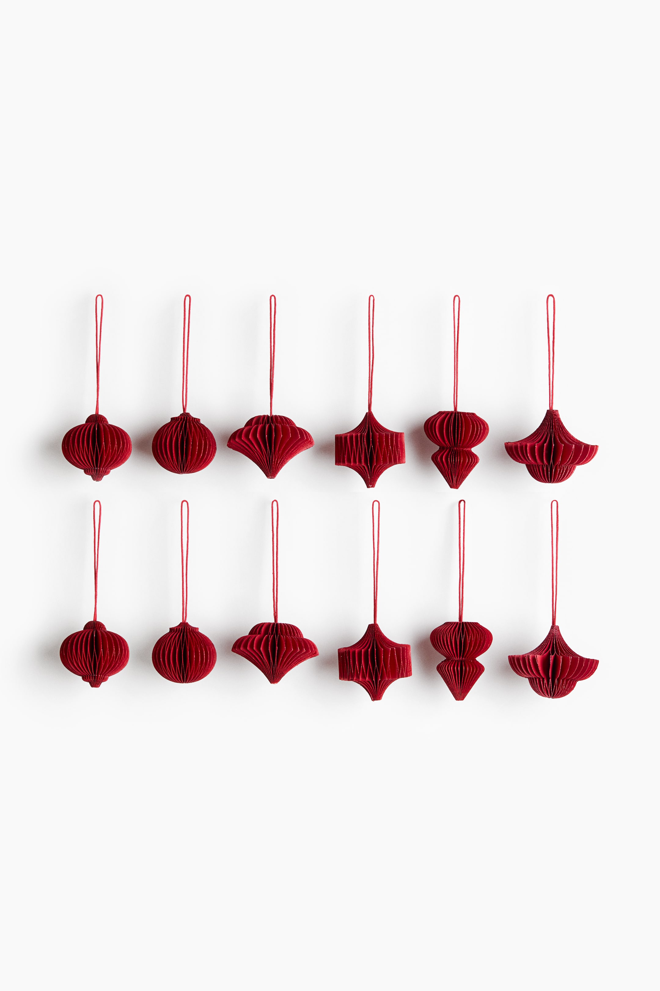 View larger image: A set of twelve deep red paper ornaments with a pleated honeycomb structure is hanging in two rows of six, shown from the front. Each ornament displays a distinct geometric shape and features a matching red string loop at the top.