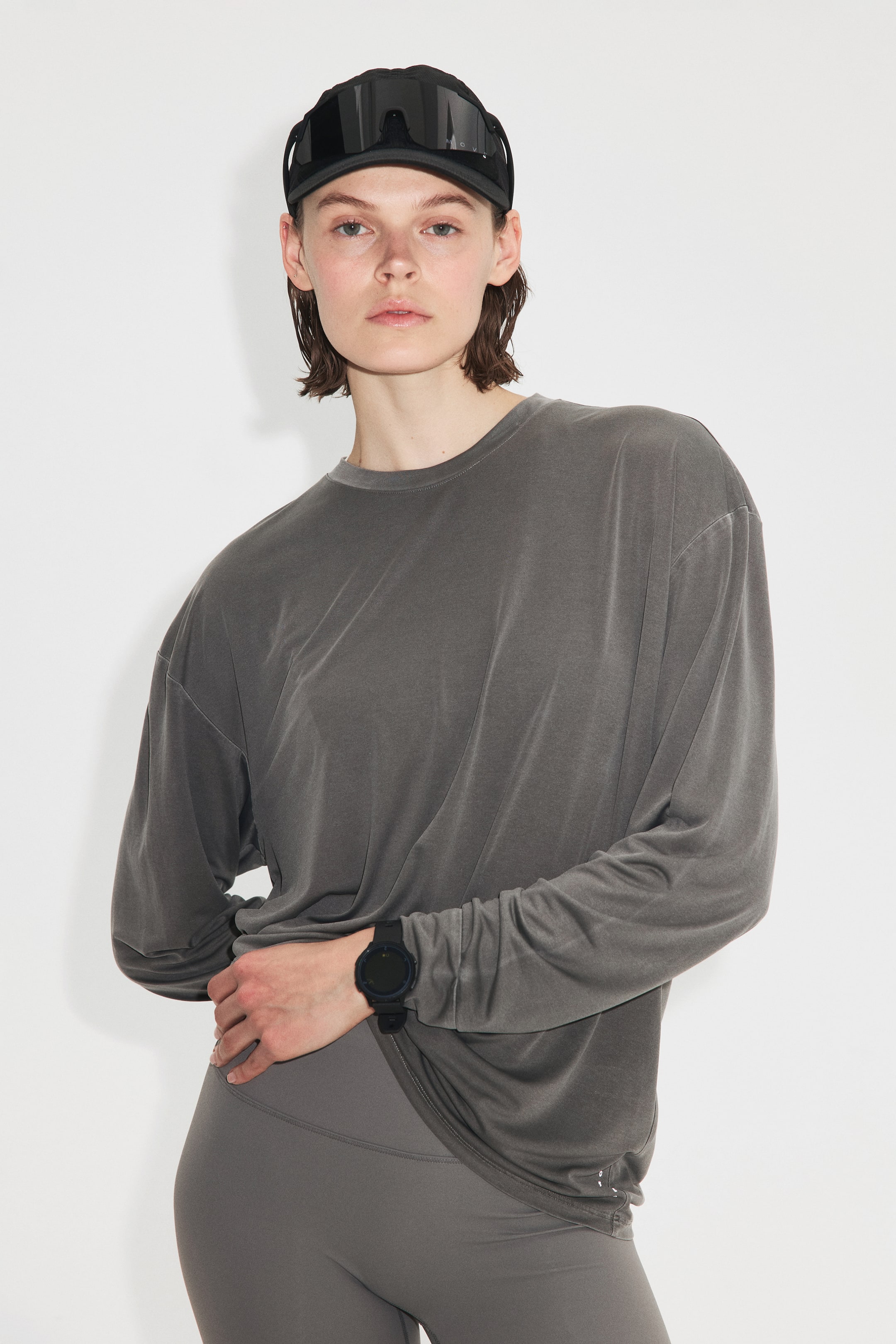 Voir une image plus grande: Un t-shirt à manches longues de couleur gris foncé délavé, à col rond, avec une coupe ample et surdimensionnée, et des manches resserrées aux poignets, est porté avec des leggings taille haute gris foncé. Une visière noire avec un large écran réfléchissant est portée vers l'avant, et une montre numérique noire est portée au poignet gauche par-dessus la manche du t-shirt.