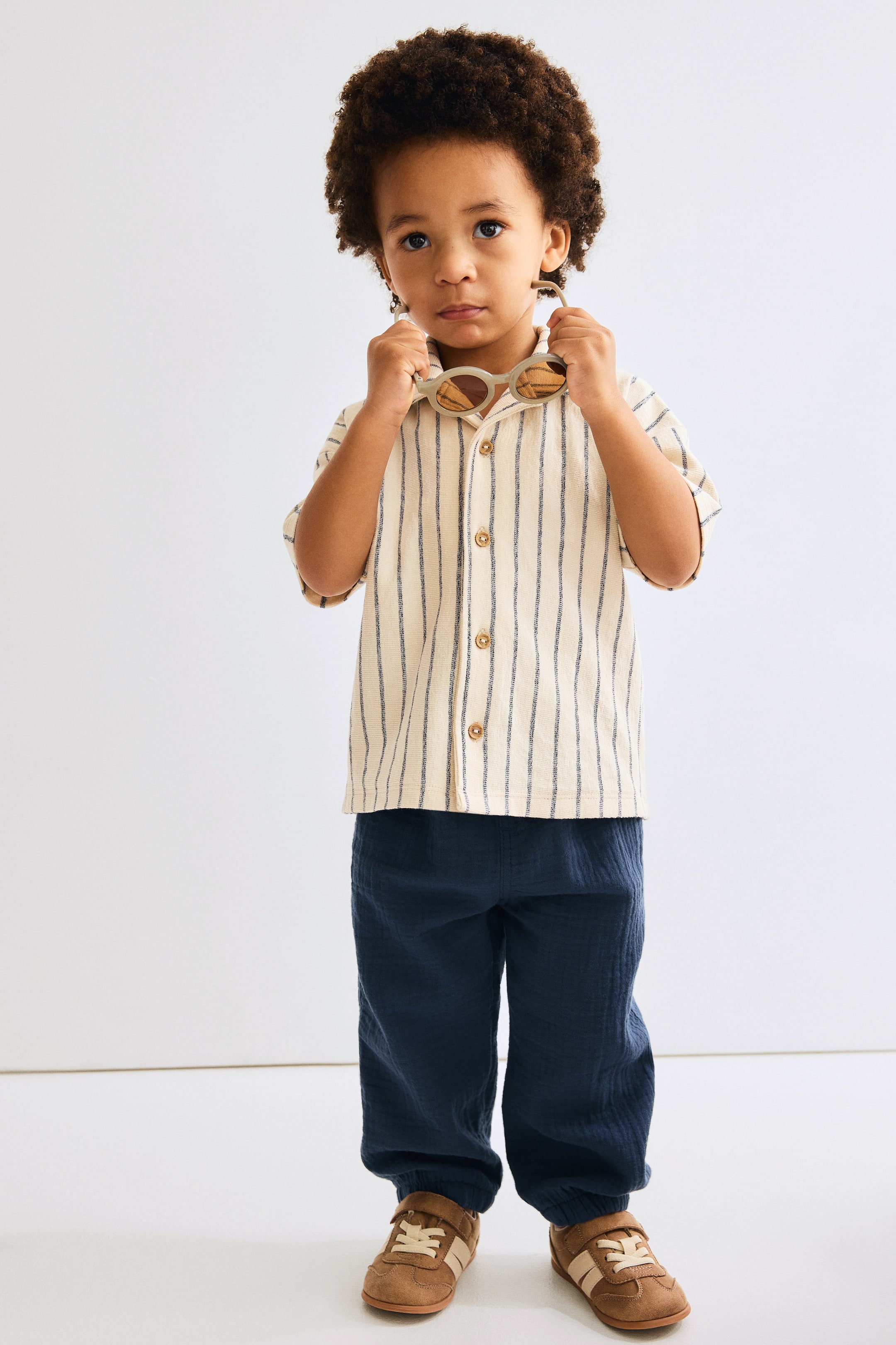 View larger image: This cream-colored, short-sleeve shirt with vertical dark stripes and a collar showcases visible buttons down the front. It is styled with crinkled dark blue pants that gather at the ankles, complemented by brown low-top sneakers featuring light stripes and Velcro closures.