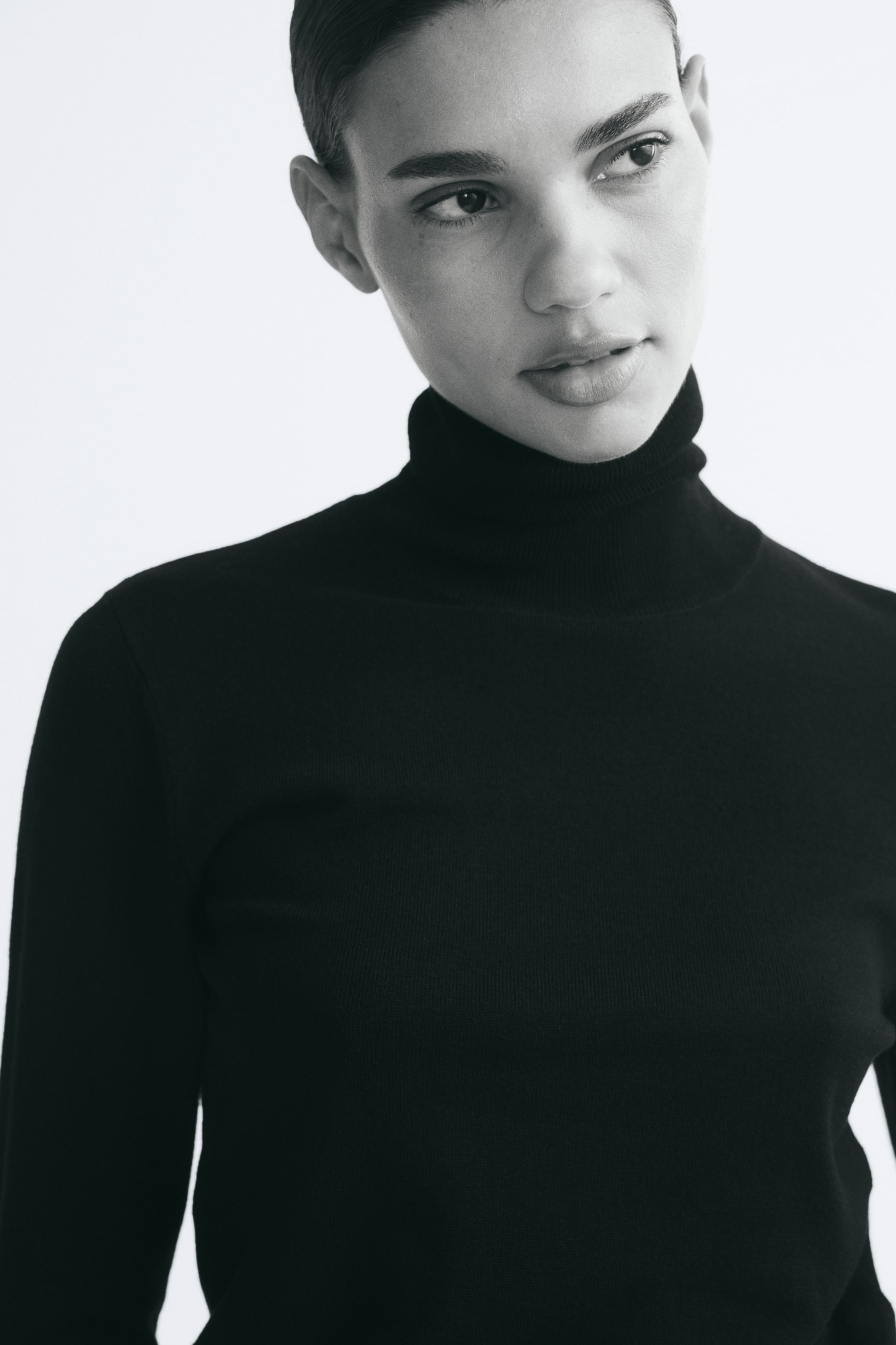 View larger image: A black, fine-knit jumper featuring a high ribbed turtle neck is being worn by the model. The close-up shot highlights the soft texture and fitted design of the long-sleeved garment.