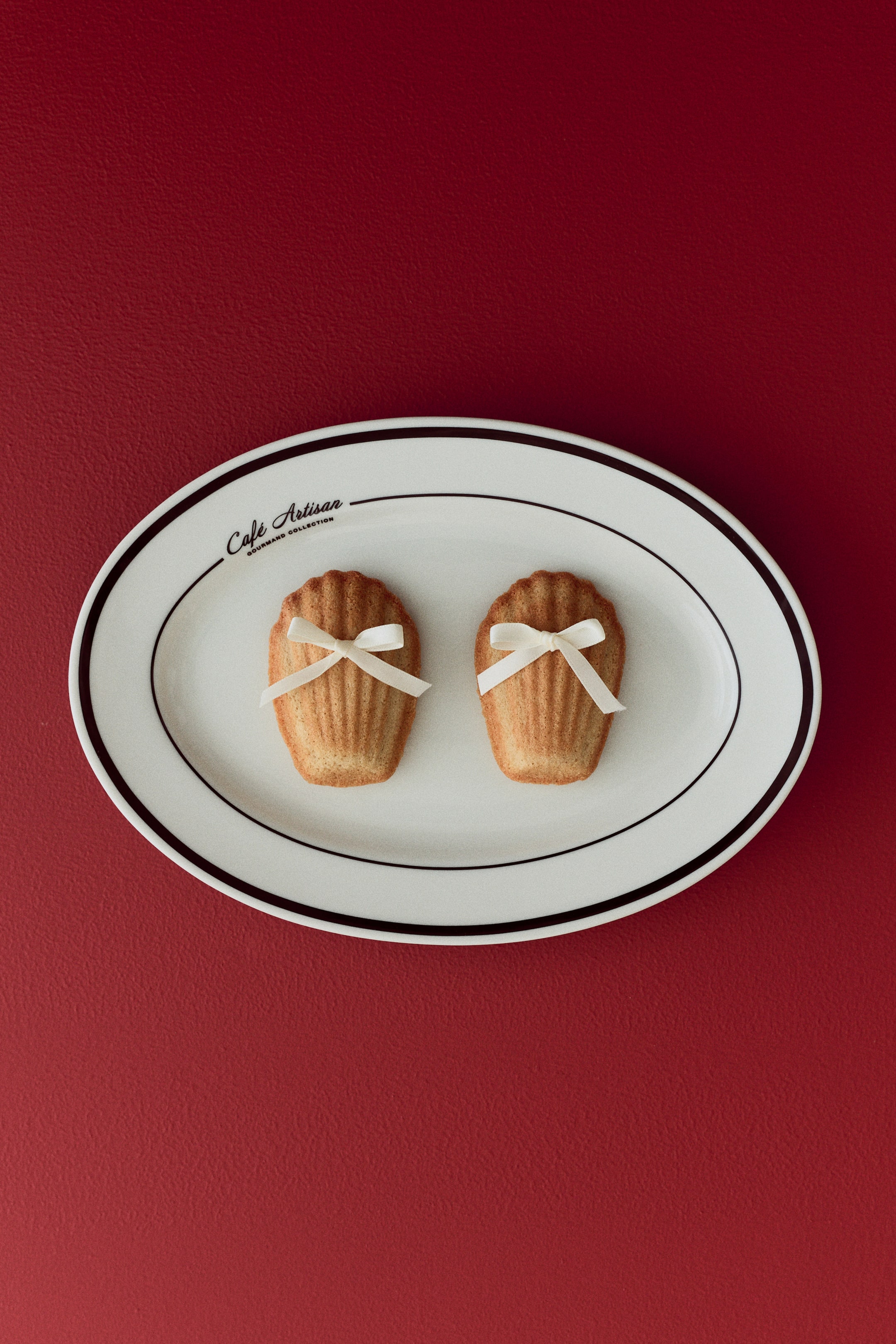 View larger image: Two golden-brown, shell-shaped madeleine cookies rest on a white oval plate, each featuring distinct ridges and a light-colored ribbon tied into a bow around the center. The oval plate features a black double-line border and the text "Cafe Artisan Gourmand Collection" at the top.