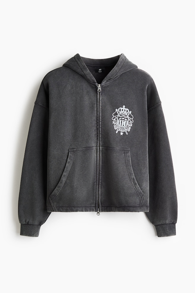 A dark grey, washed-finish zip-up hoodie is facing the front, featuring a hood, a full-length front zipper, and a split kangaroo pocket. It is displaying a white crest graphic on the left chest alongside dropped shoulders and ribbed cuffs. - 1