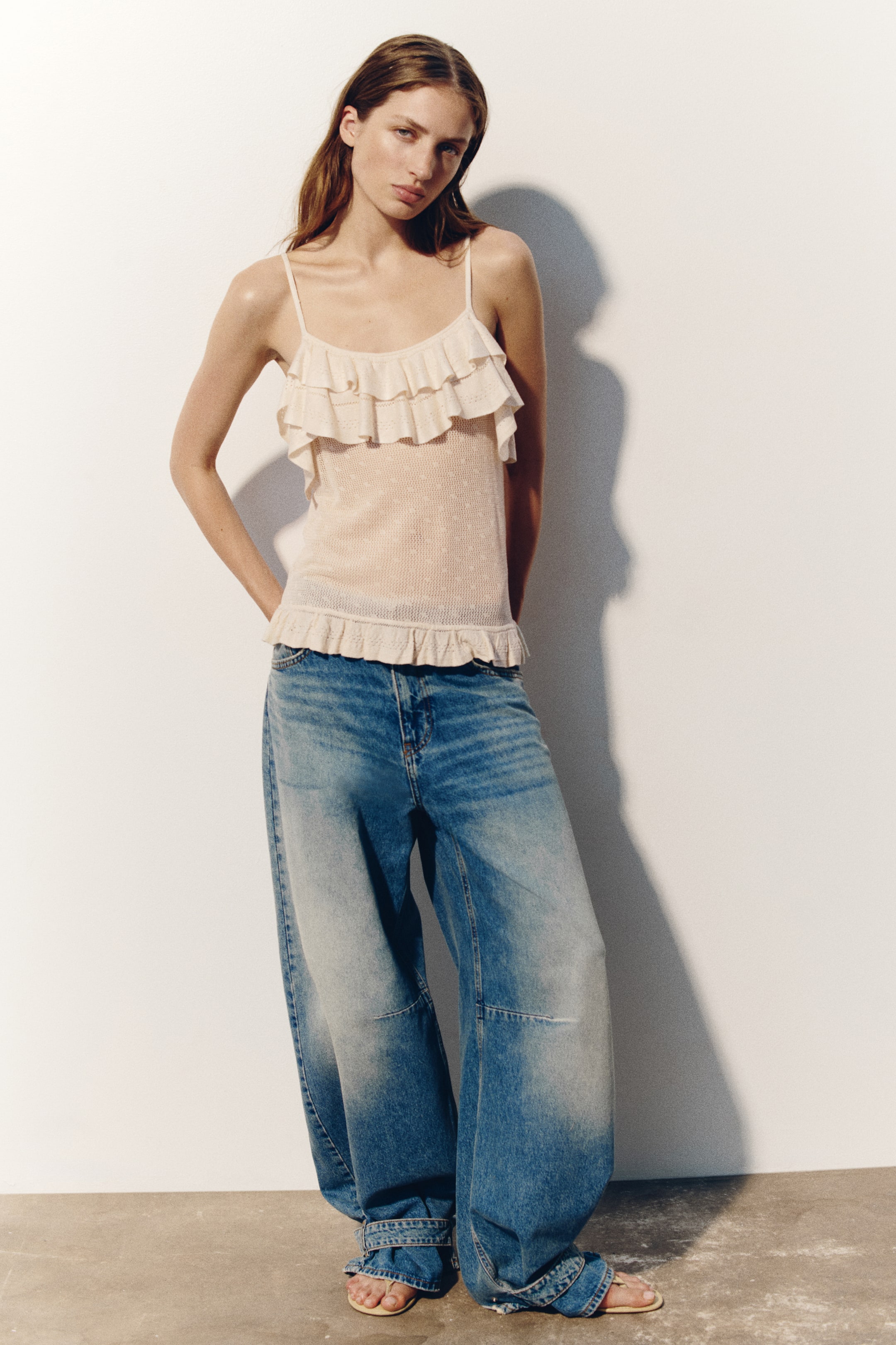 View larger image: A light-coloured top in a soft pointelle knit, featuring a flounced scoop neckline, spaghetti shoulder straps, and a flounced hem, is being worn by the model, styled with wide-leg blue jeans.