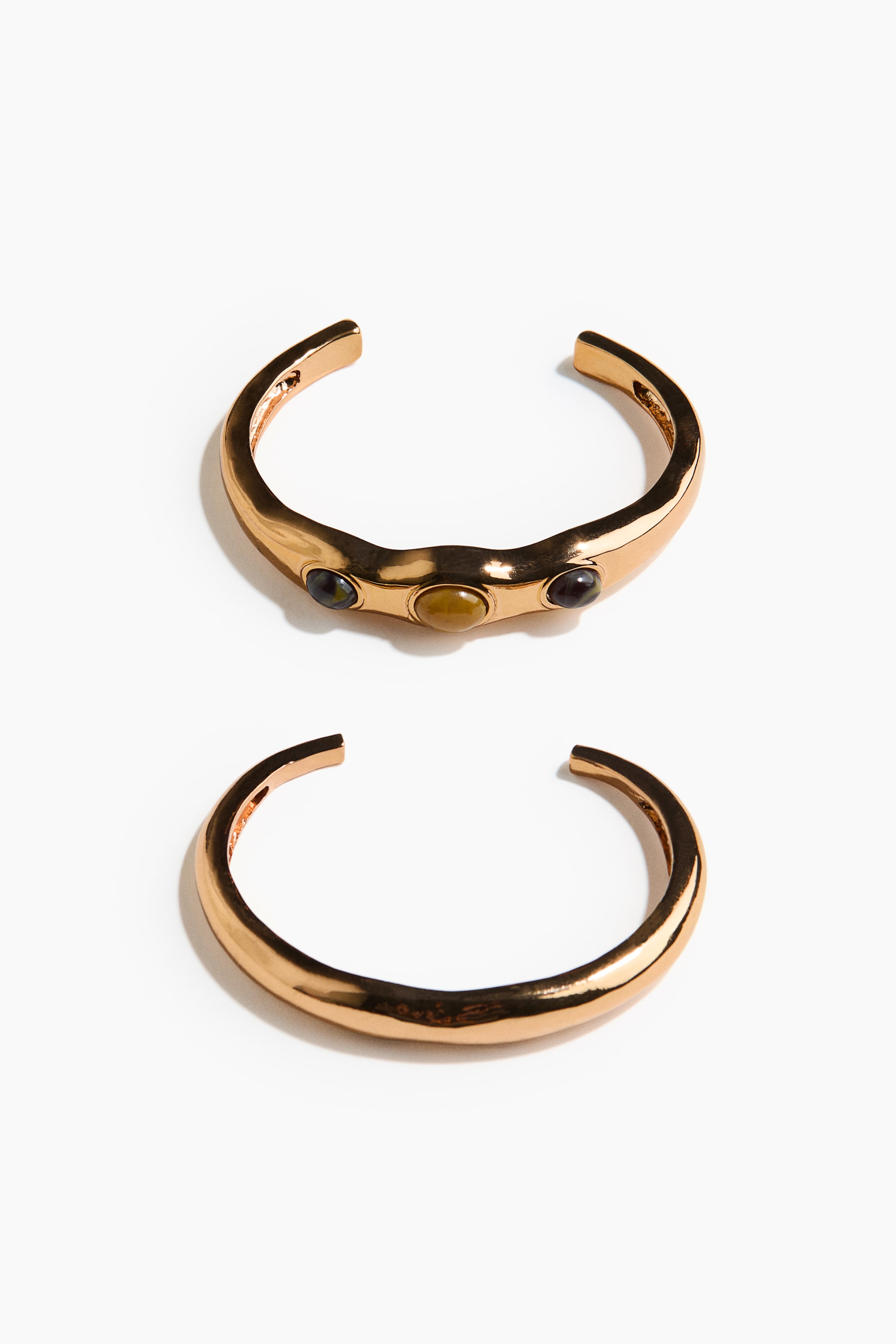 View larger image: Two gold-toned open cuff bracelets are displayed. The upper cuff is featuring three oval cabochon stones in dark green, amber, and dark purple, and is exhibiting an organic, textured surface. The lower cuff is smooth and polished.