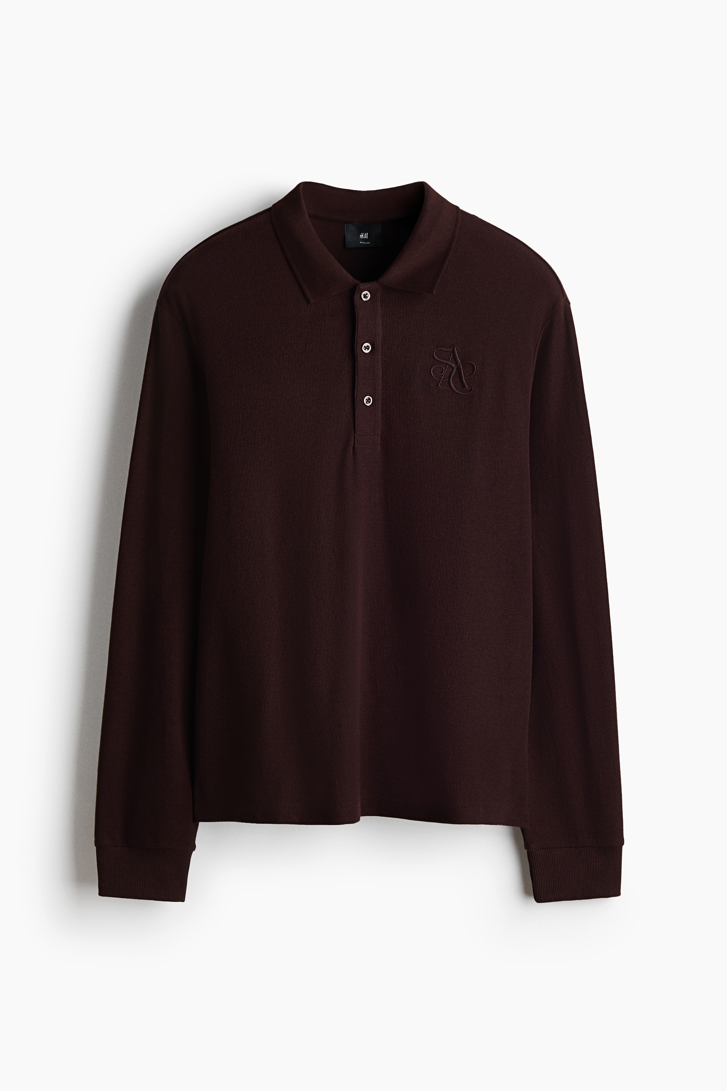 A dark brown, long-sleeved polo shirt in waffle-knit fabric is being worn by the model. The shirt features a ribbed collar, a button placket, and a tonal embroidered detail on the chest, and is paired with off-white trousers.