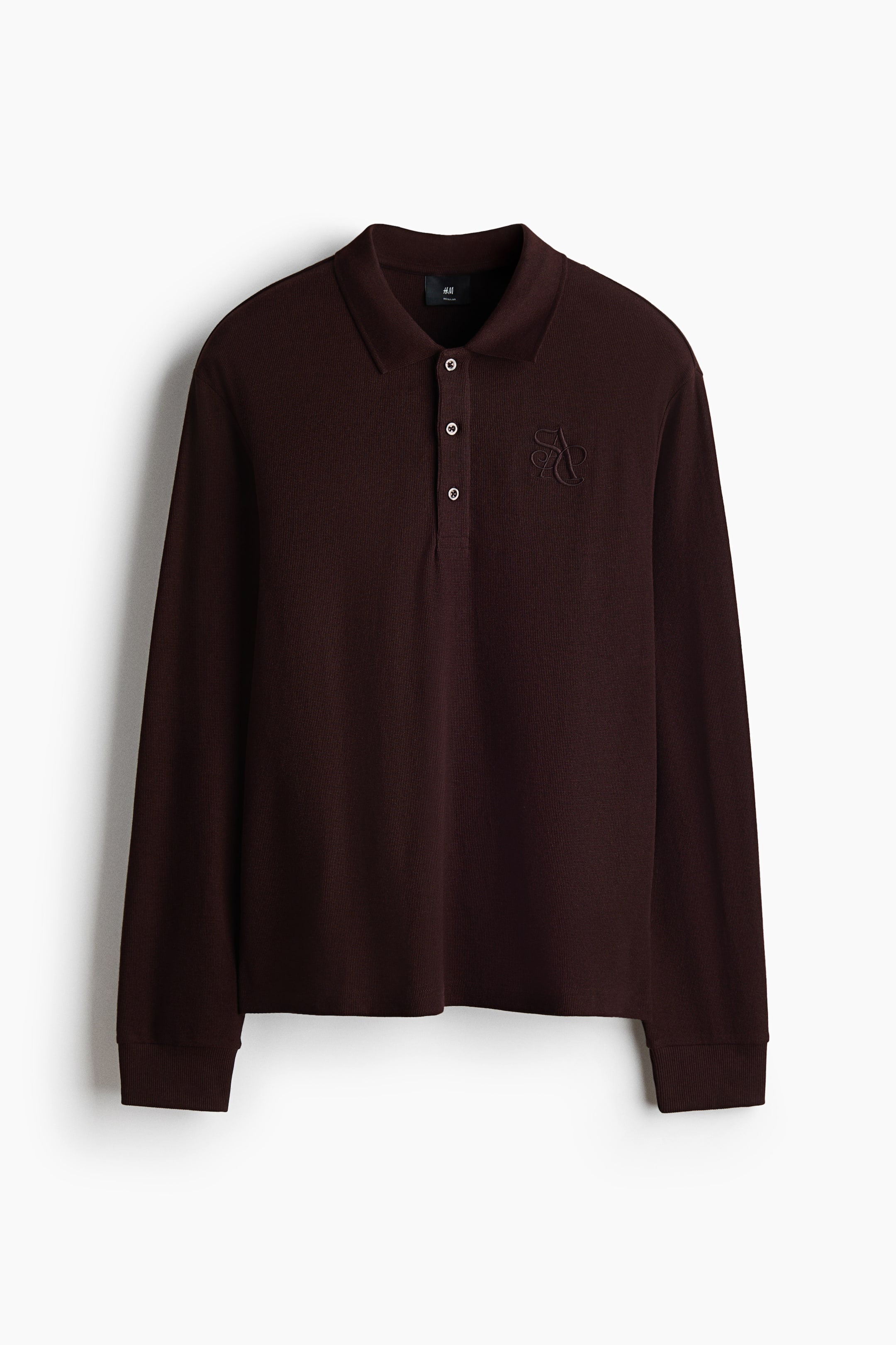 View larger image: A dark brown long-sleeved polo shirt is showcasing its front view. It features a collar and a three-button placket, with light-colored buttons fastening the opening. An embroidered monogram matching the shirt color adorns the left chest, and ribbed cuffs complete the long sleeves.