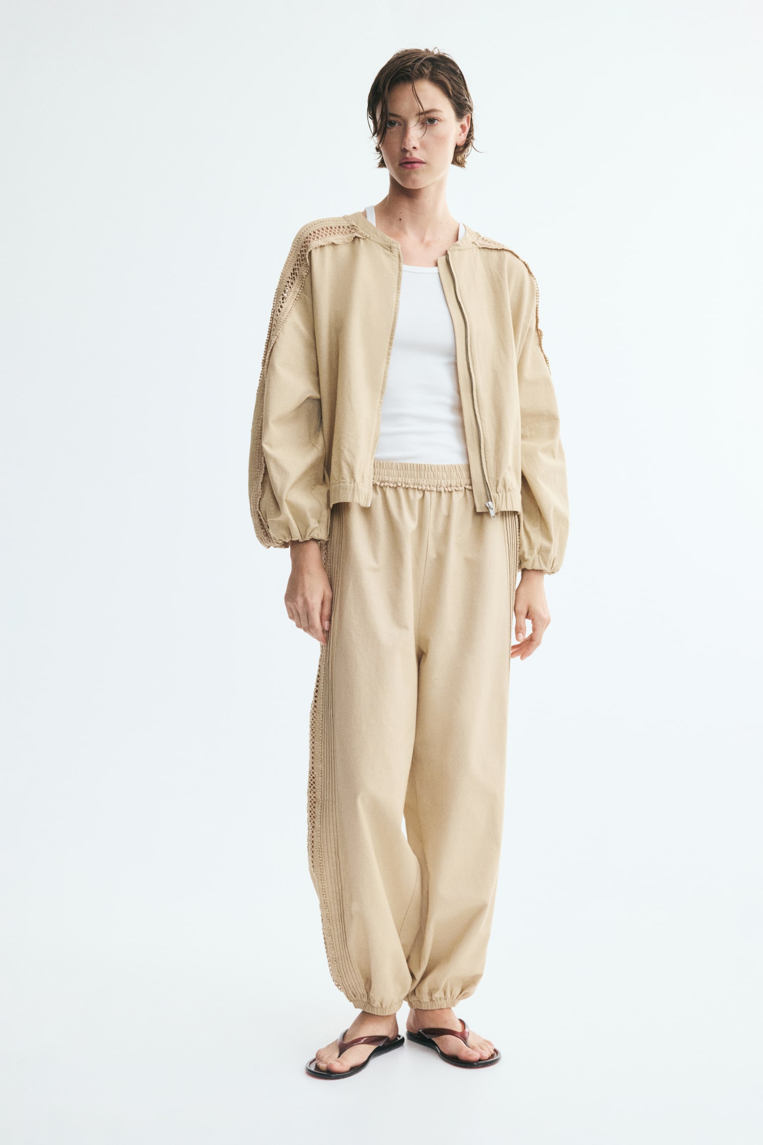 Beige trousers are showcasing a shirred waistband and shirred ankle cuffs. Vertical panels run along the outer seams, featuring multiple pin tucks and an openwork geometric lace pattern. - 1