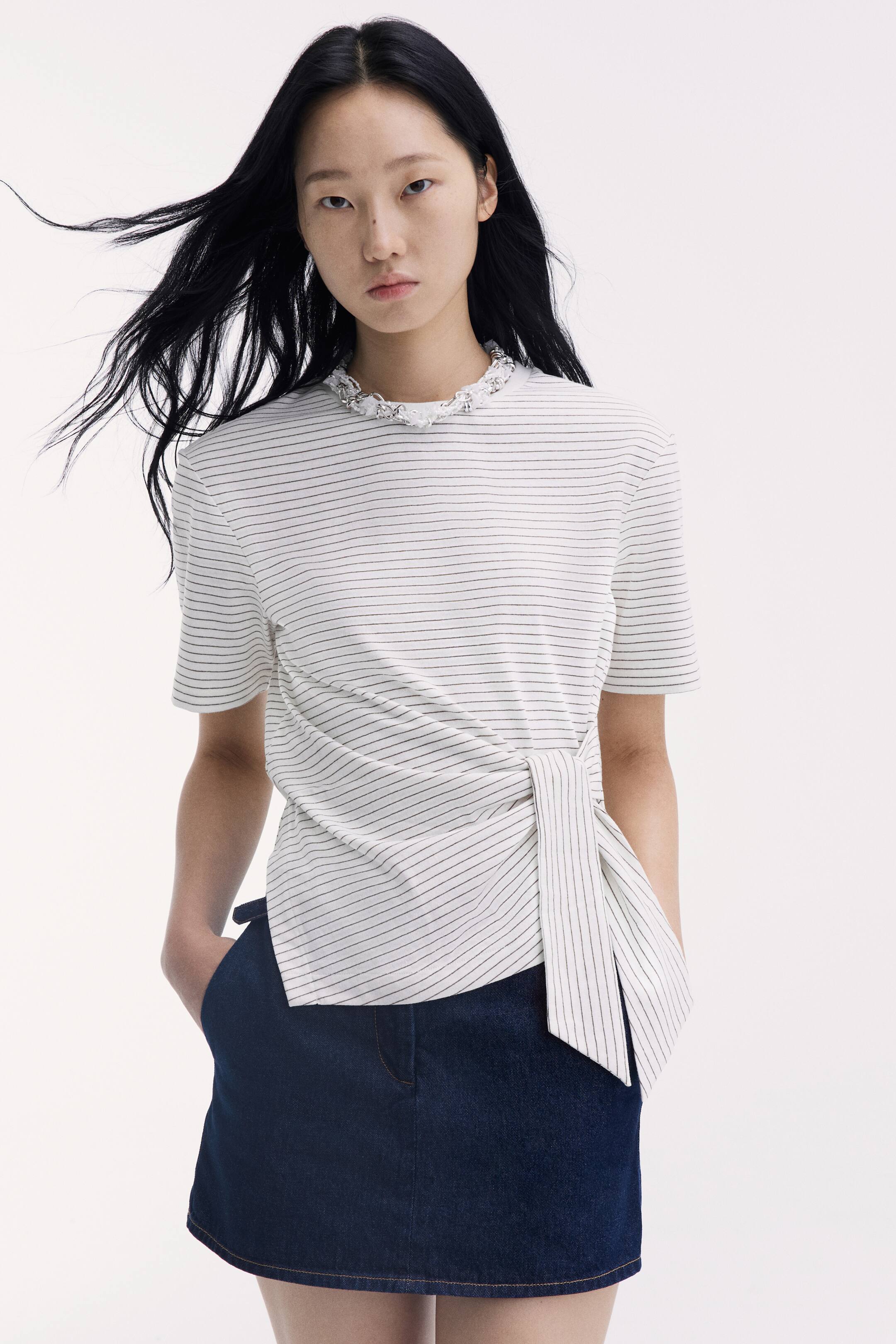 View larger image: The model is wearing a short dark blue denim skirt, with one hand placed in its diagonal side pocket. The skirt is styled with a white-and-black striped short-sleeved top that drapes over the waistband.