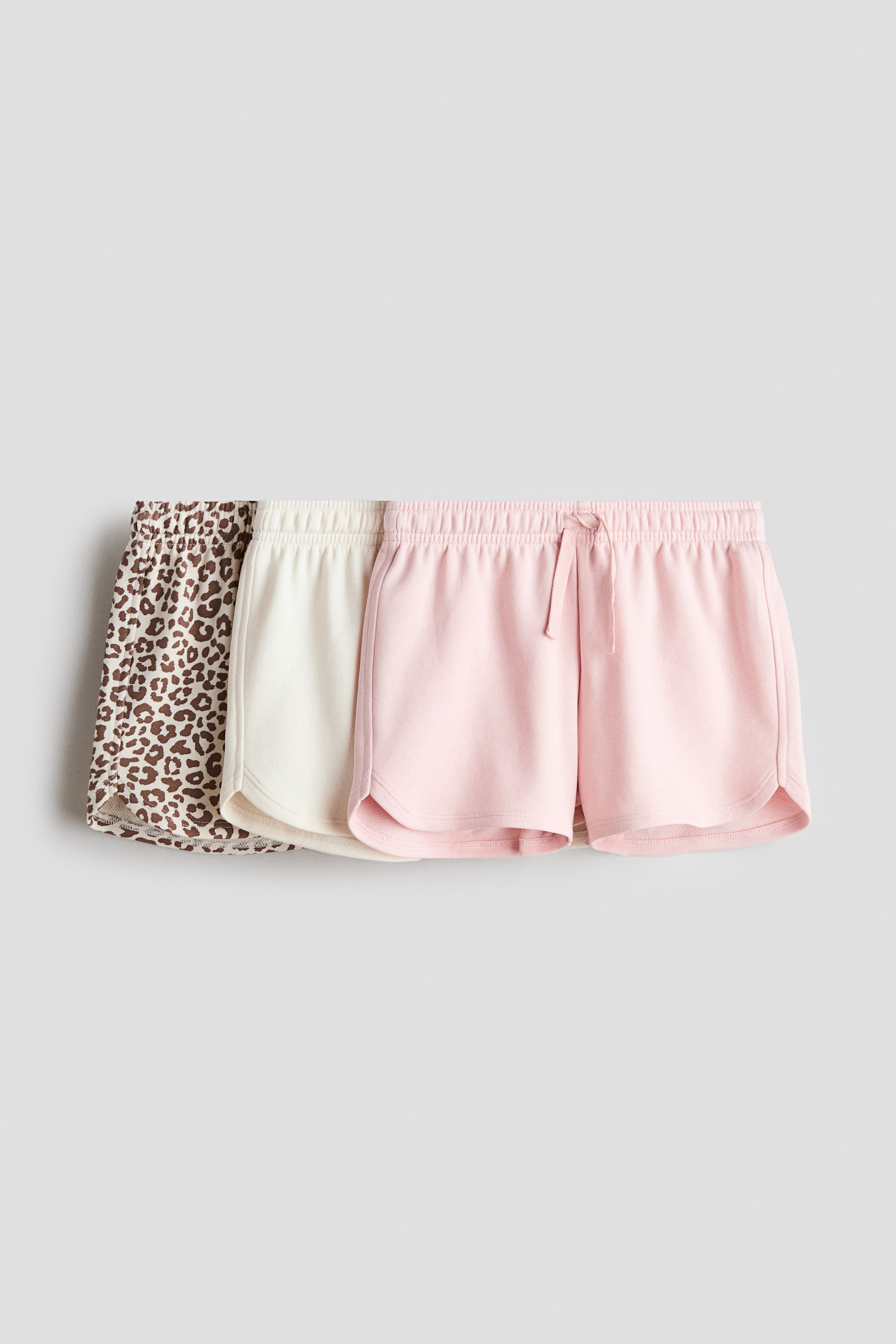 View larger image: Three pairs of shorts are layered, with the front-facing light pink pair featuring an elasticated waistband with a drawstring and curved hems. A cream pair is layered beneath it, and a brown and black leopard print pair is visible at the bottom, both also showing elasticated waistbands and curved hems.