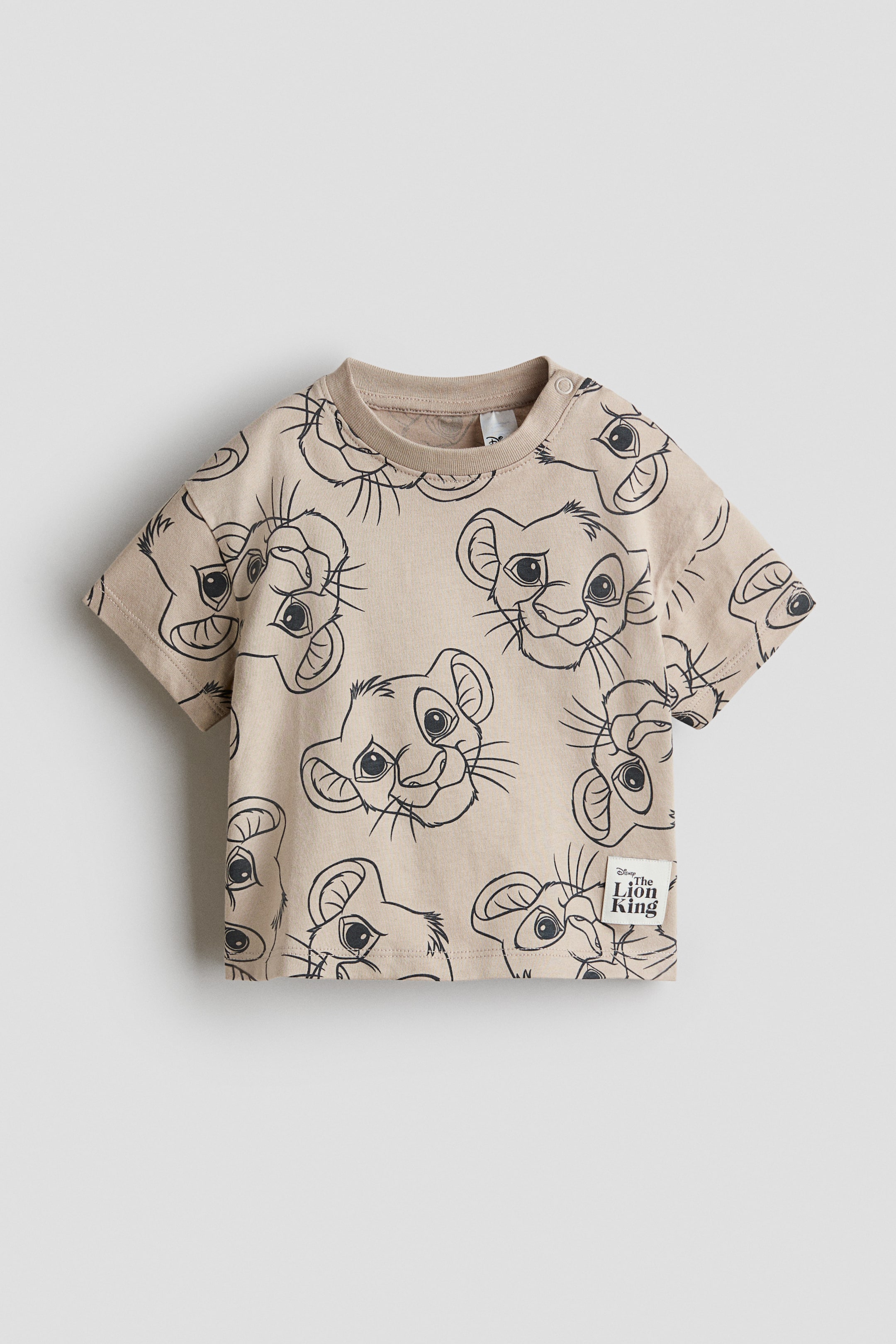View larger image: Printed cotton T-shirt. Displayed from the front, it showcases an allover print of black outline cartoon lion cub faces on a beige background. Designed with a ribbed crew neck, short sleeves, two snap buttons visible on the wearer's right shoulder, and a "Disney The Lion King" fabric label attached to the bottom right hem.