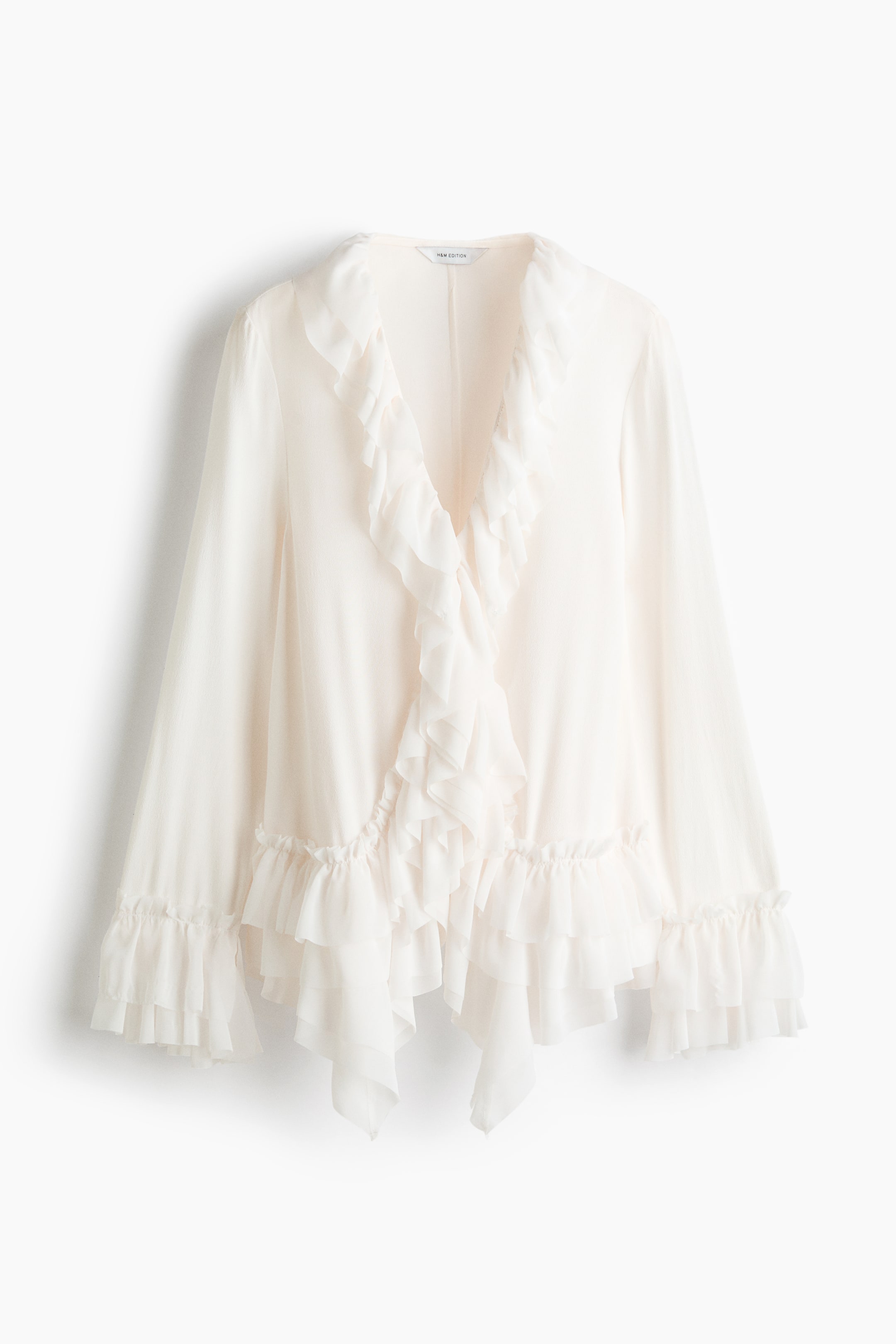 Ver imagem maior: Uma blusa longa, em tom creme, em tecido de viscose leve, é apresentada de frente. Apresenta um decote em V e fecho frontal oculto com colchetes. Camadas de folhos volumosos decoram o decote, prolongam-se pela frente e adornam os punhos e a bainha.