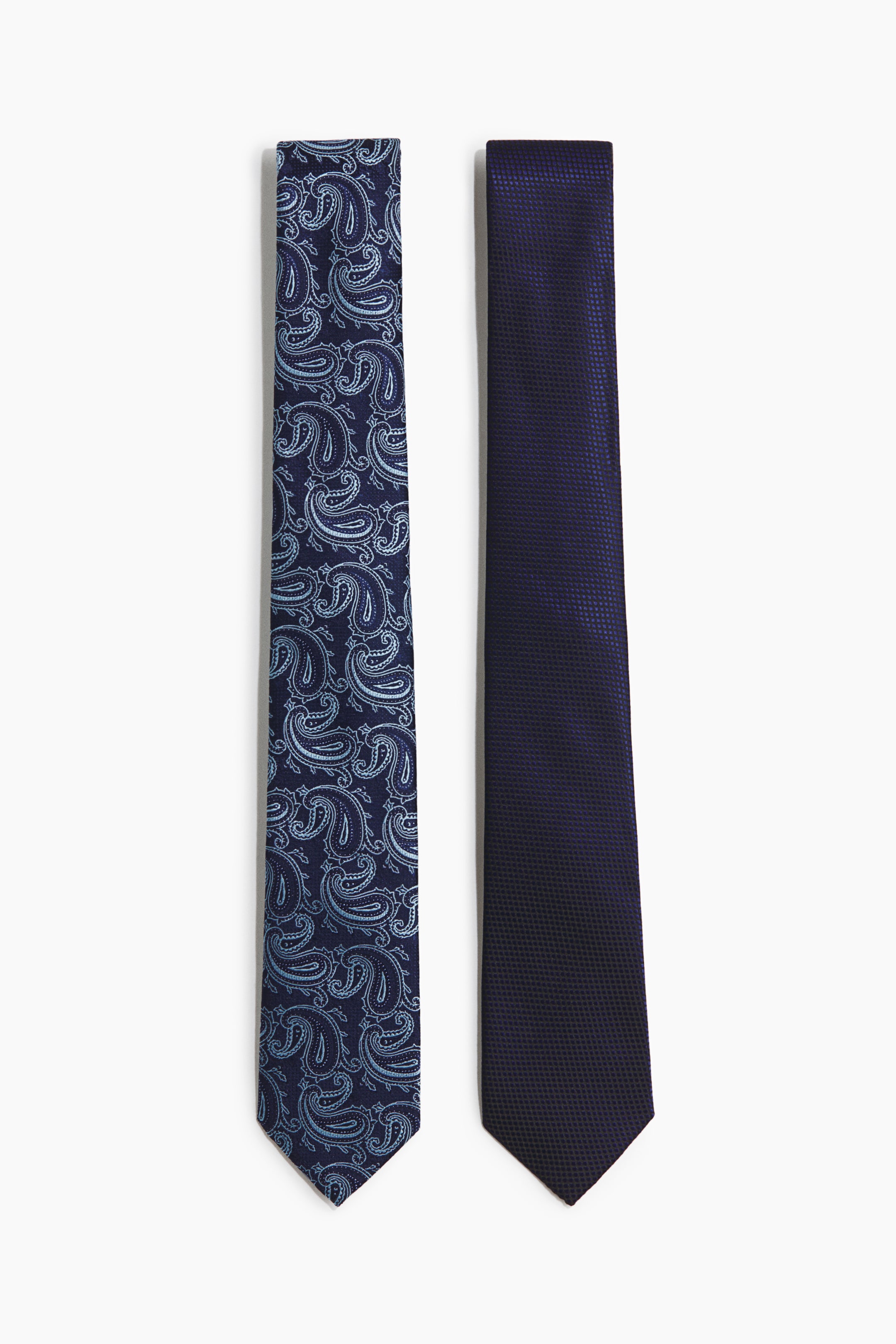 Ampliar la imagen: Dos corbatas clásicas están colocadas planas, mirando hacia adelante. La corbata de la izquierda presenta una tela tejida de color azul oscuro con un patrón de paisley en azul claro, y la corbata de la derecha muestra una tela tejida de color azul oscuro con un patrón texturizado sutil.