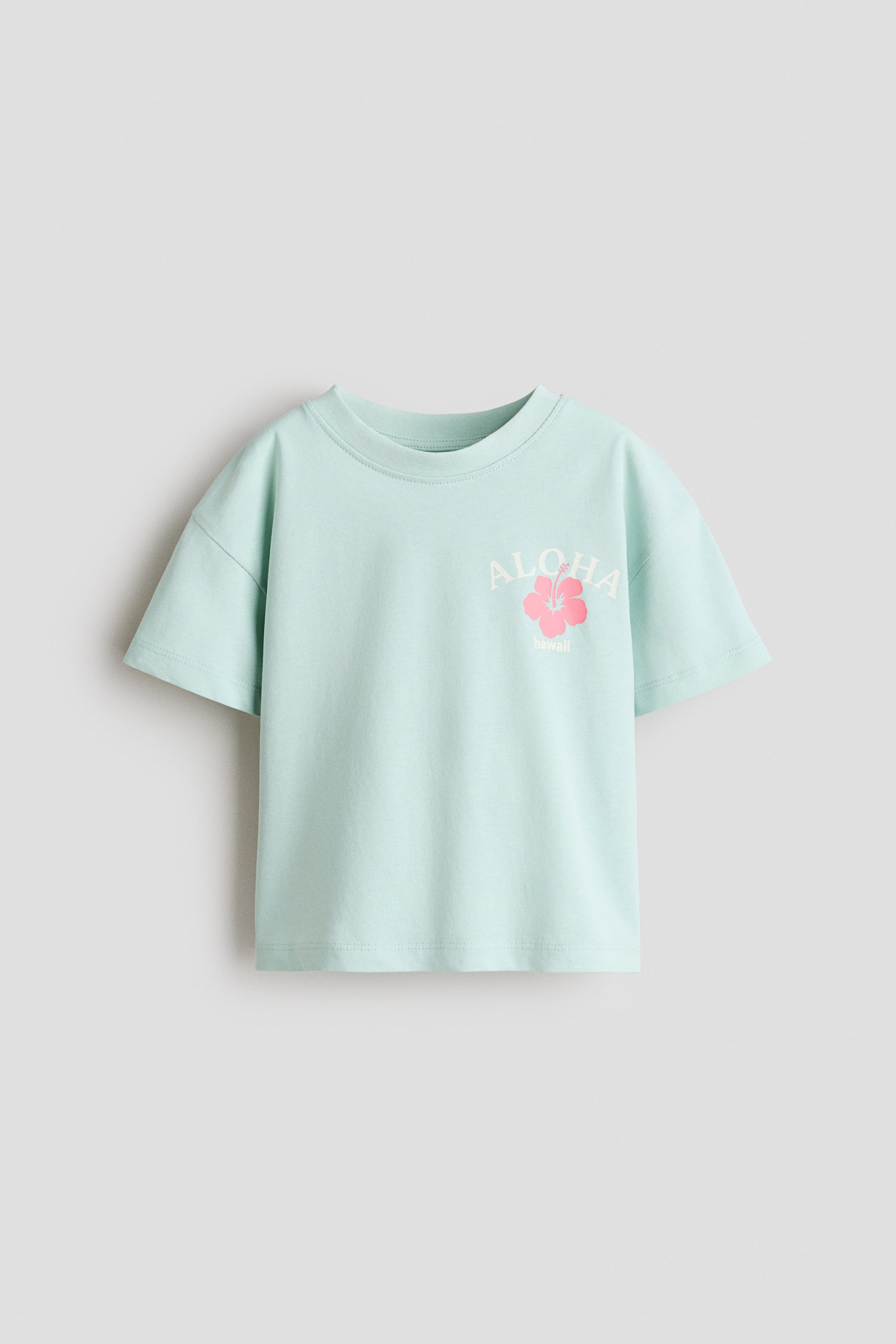 拡大画像を表示: プリントコットンジャージー素材のライトブルーのリラックスフィットTシャツがフロントに登場。丸いリブトリムのネックライン、ドロップショルダー、胸元には白の「ALOHA Hawaii」グラフィックとピンクのハイビスカスの花が特徴です。