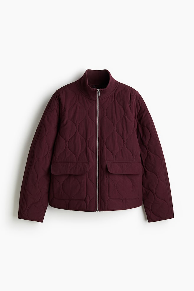 A dark red quilted jacket is presented front-facing, featuring a scalloped quilting pattern. It is designed with a ribbed stand collar, a silver-toned front zipper, and two large flap pockets on the lower front. - 1