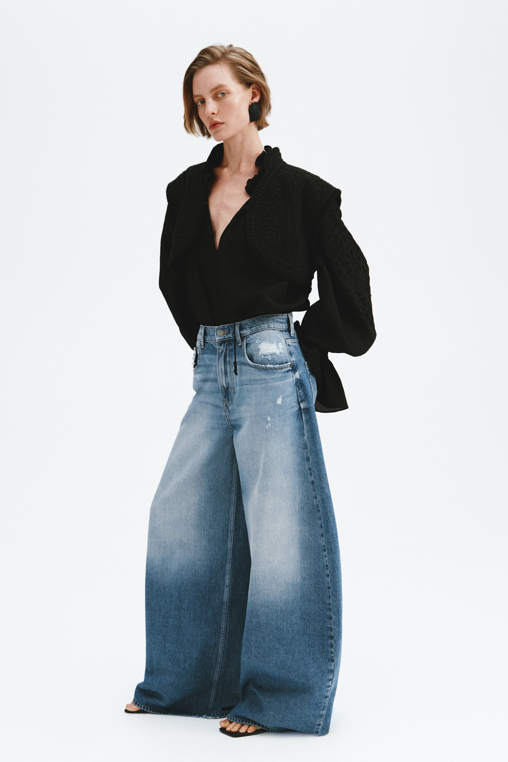 Grotere afbeelding bekijken: Een zwarte blouse met een diepe V-hals en een gerimpelde opstaande kraag, voorzien van volumineuze mouwen en een textuurdetail op de schouders. Deze wordt gecombineerd met een hoog getailleerde, wijde blauwe jeans met een licht-naar-donker ombre wassing en versleten accenten.