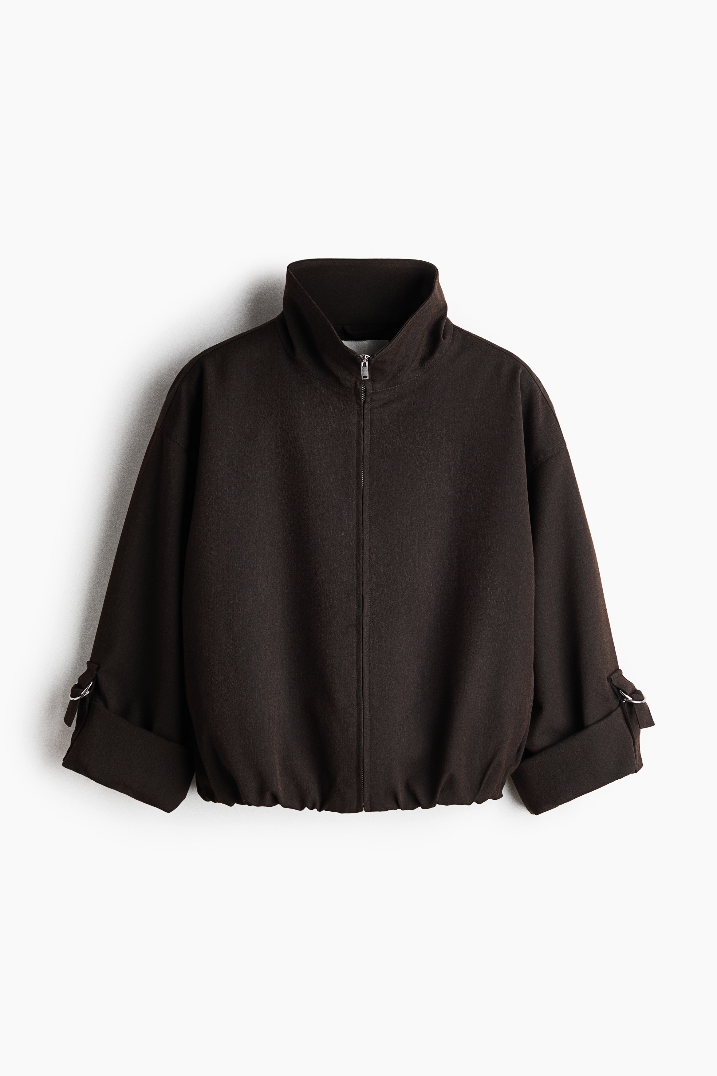 Das Model trÃ¤gt eine kurz geschnittene, oversized Jacke in Dunkelbraun mit Stehkragen und teilweise geÃ¶ffnetem ReiÃverschluss, Ã¼berschnittenen Schultern und umgeschlagenen Manschetten, die mit Metall-D-Ringen befestigt sind. Die Jacke wird Ã¼ber einem schwarzen Oberteil mit hohem Kragen und schwarzen Hosen getragen.
