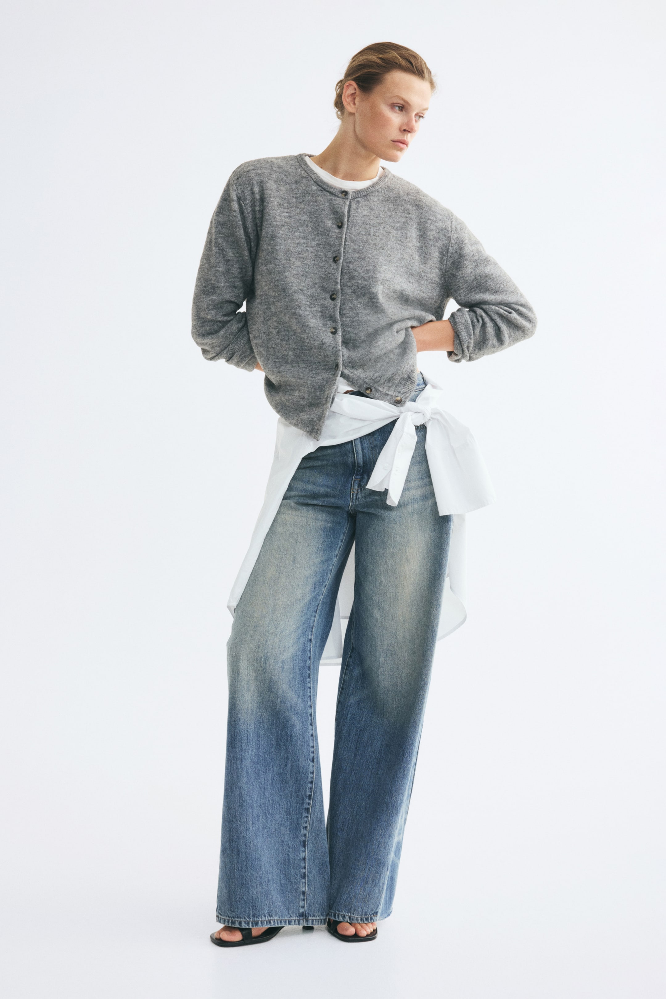View larger image: A grey, soft-knit cardigan featuring a round neckline, a button-front closure, and ribbed trims is worn by the model. The cardigan is layered over a white crew-neck T-shirt and paired with wide-leg blue jeans and a white shirt tied around the waist.