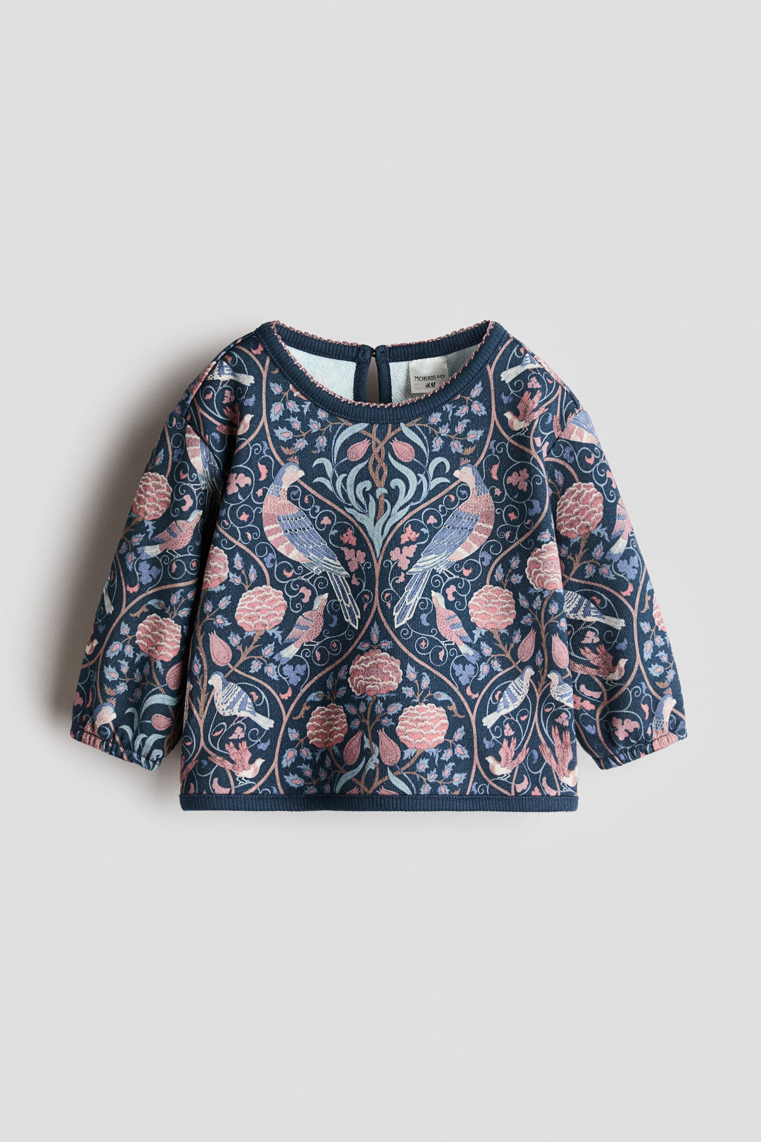 Egy kÃ¶nnyÅ±, pamutbÃ³l kÃ©szÃ¼lt sweatshirt lÃ¡thatÃ³ elÃ¶lnÃ©zetbÅl, teljes felÃ¼letÃ©n szimmetrikus, madarakat Ã©s virÃ¡gmintÃ¡kat Ã¡brÃ¡zolÃ³, rÃ³zsaszÃ­n Ã©s kÃ©k Ã¡rnyalatÃº nyomattal, sÃ¶tÃ©t tengerÃ©szkÃ©k alapon. A ruhadarabon leejtett vÃ¡llmegoldÃ¡s, keskeny elasztikus mandzsettÃ¡val ellÃ¡tott hosszÃº ujjak, kerek, picot szegÃ©lyÅ± nyakkivÃ¡gÃ¡s, valamint bordÃ¡zott szegÃ©lyÅ± alsÃ³ rÃ©sz lÃ¡thatÃ³. - 1